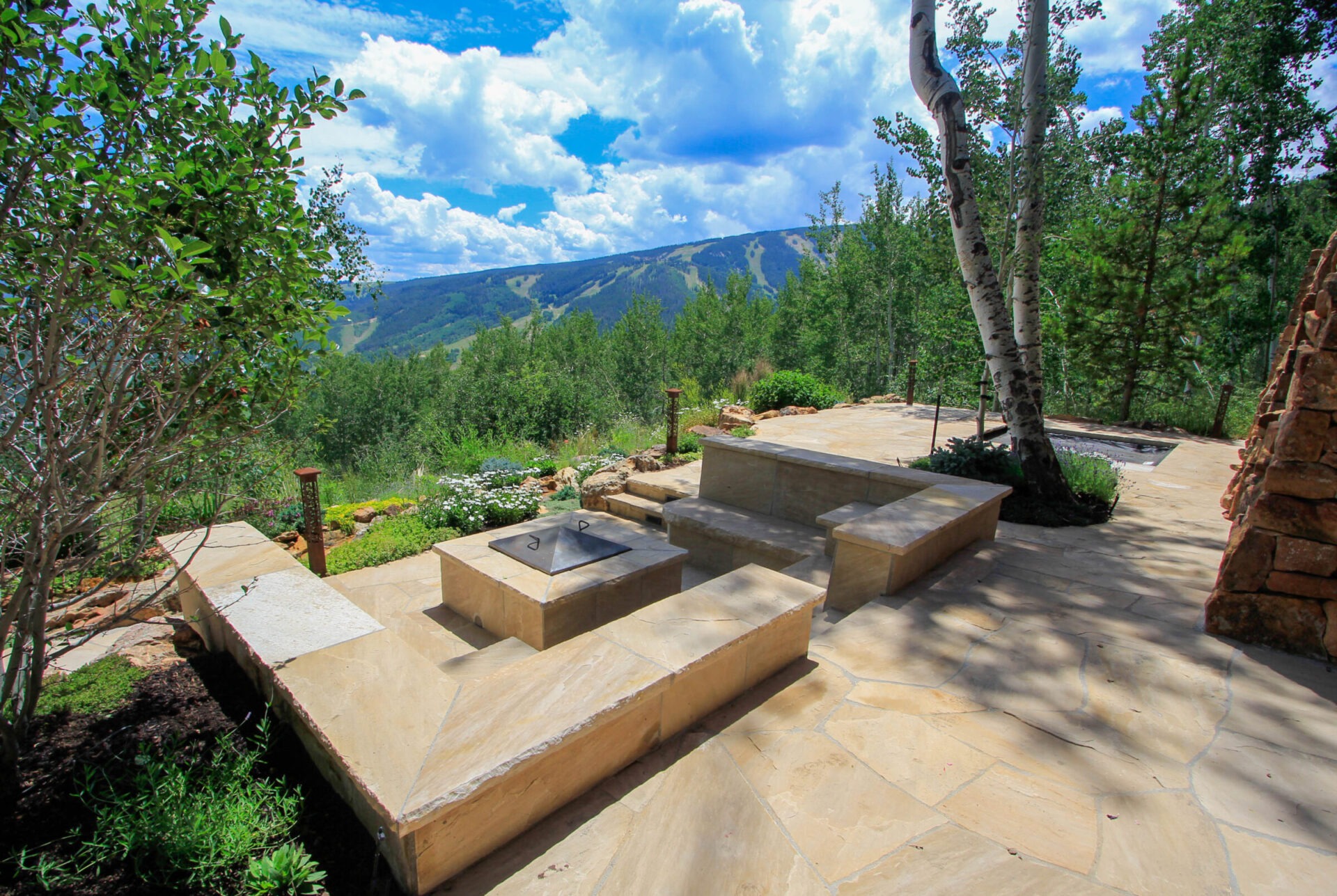 An outdoor stone patio with benches, a fire pit, lush greenery, and mountain views under a blue sky with fluffy clouds.