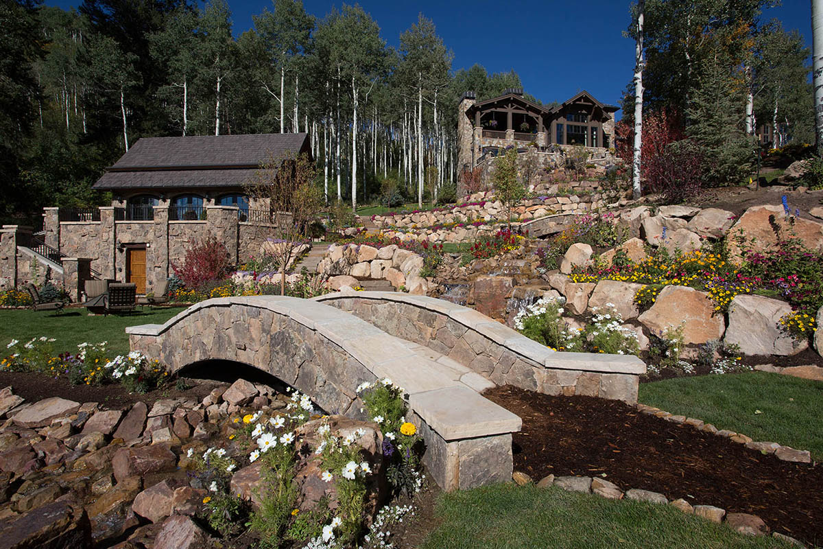 A luxurious house with large windows is surrounded by a landscaped garden, stone walls, colorful flowers, a small stone bridge, and a backdrop of aspen trees.