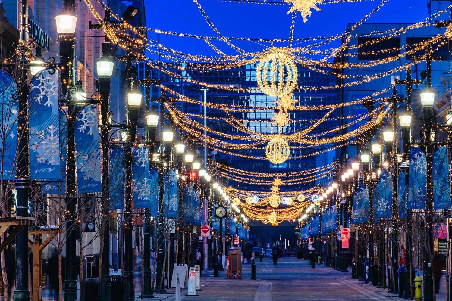 An urban street adorned with festive lights and decorations in the twilight. Illuminated street lamps and ornate light fixtures create a holiday atmosphere.