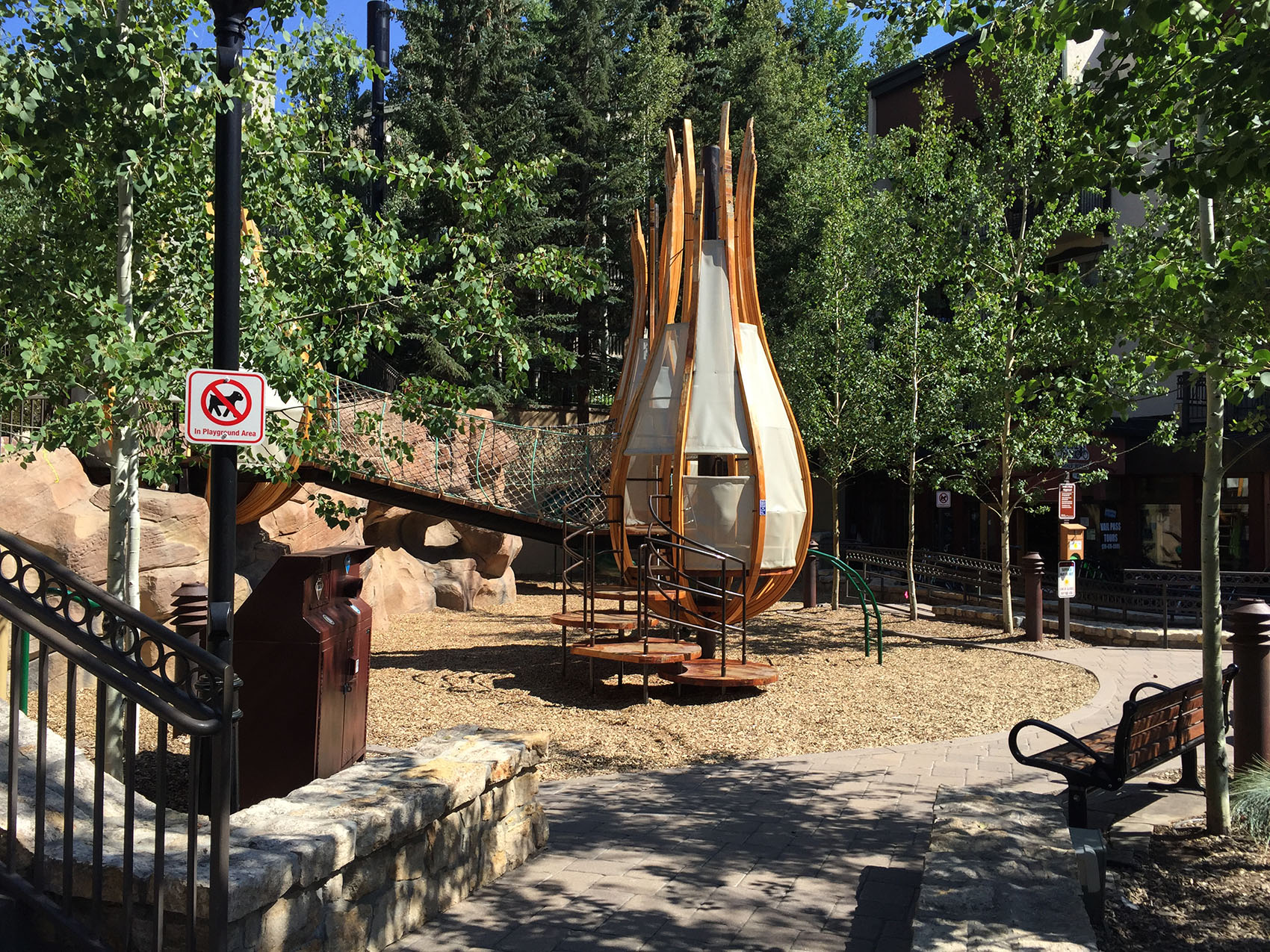 An outdoor playground mimicking a teepee structure, surrounded by trees, with a no smoking sign and benches under a clear sunny sky.