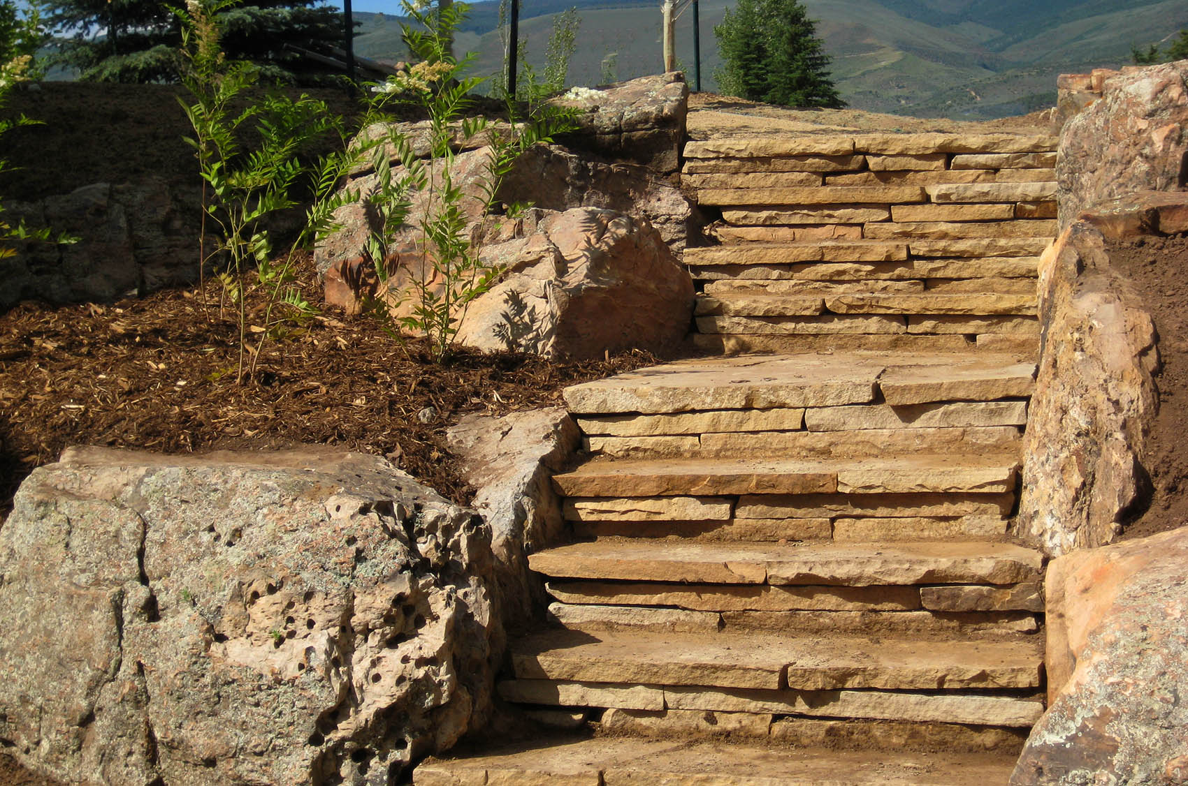 Natural stone steps ascend between large boulders and freshly mulched garden beds, with mountainous landscape and clear sky in the background.