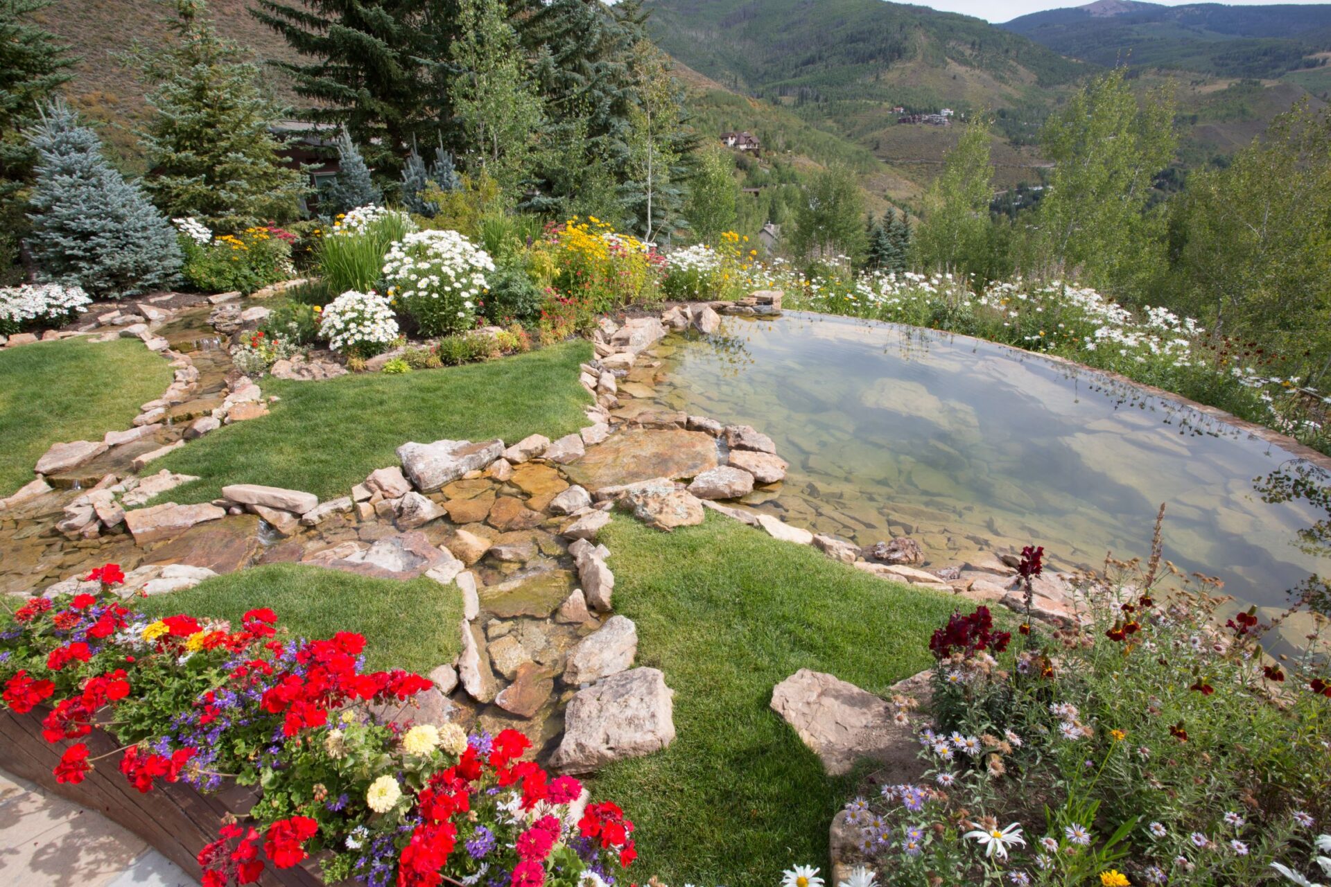 A serene garden with a natural stone-edged pond surrounded by lush greenery, colorful flowers, and a hilly landscape in the background.