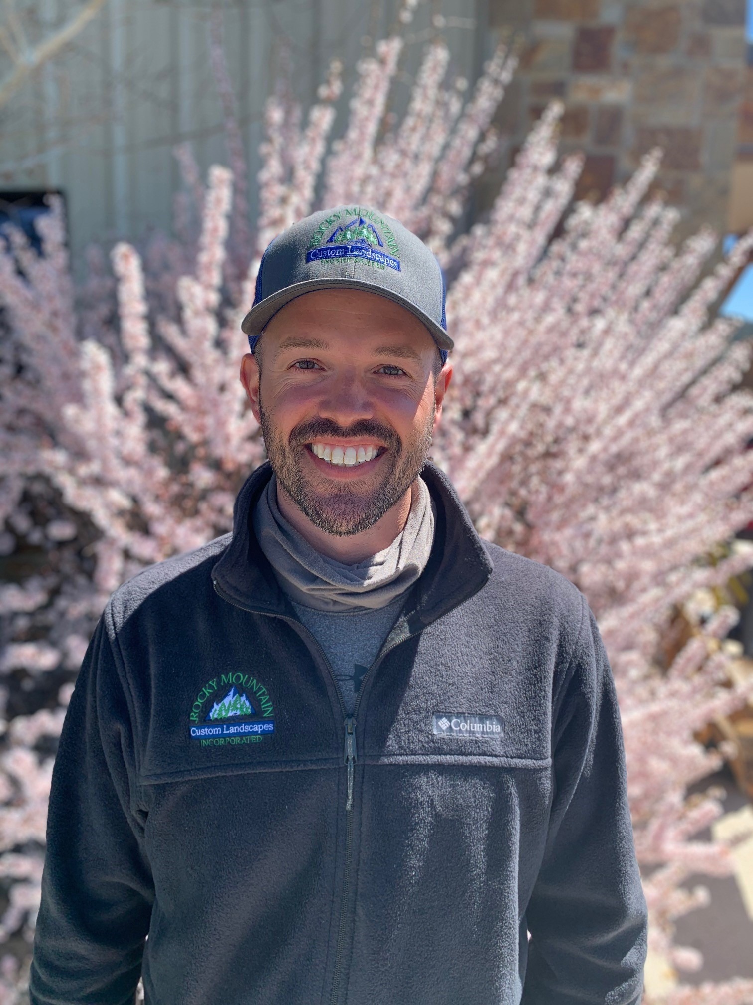 A person with a beard and cap is smiling in front of blooming pink flowers, wearing a fleece jacket on a sunny day.
