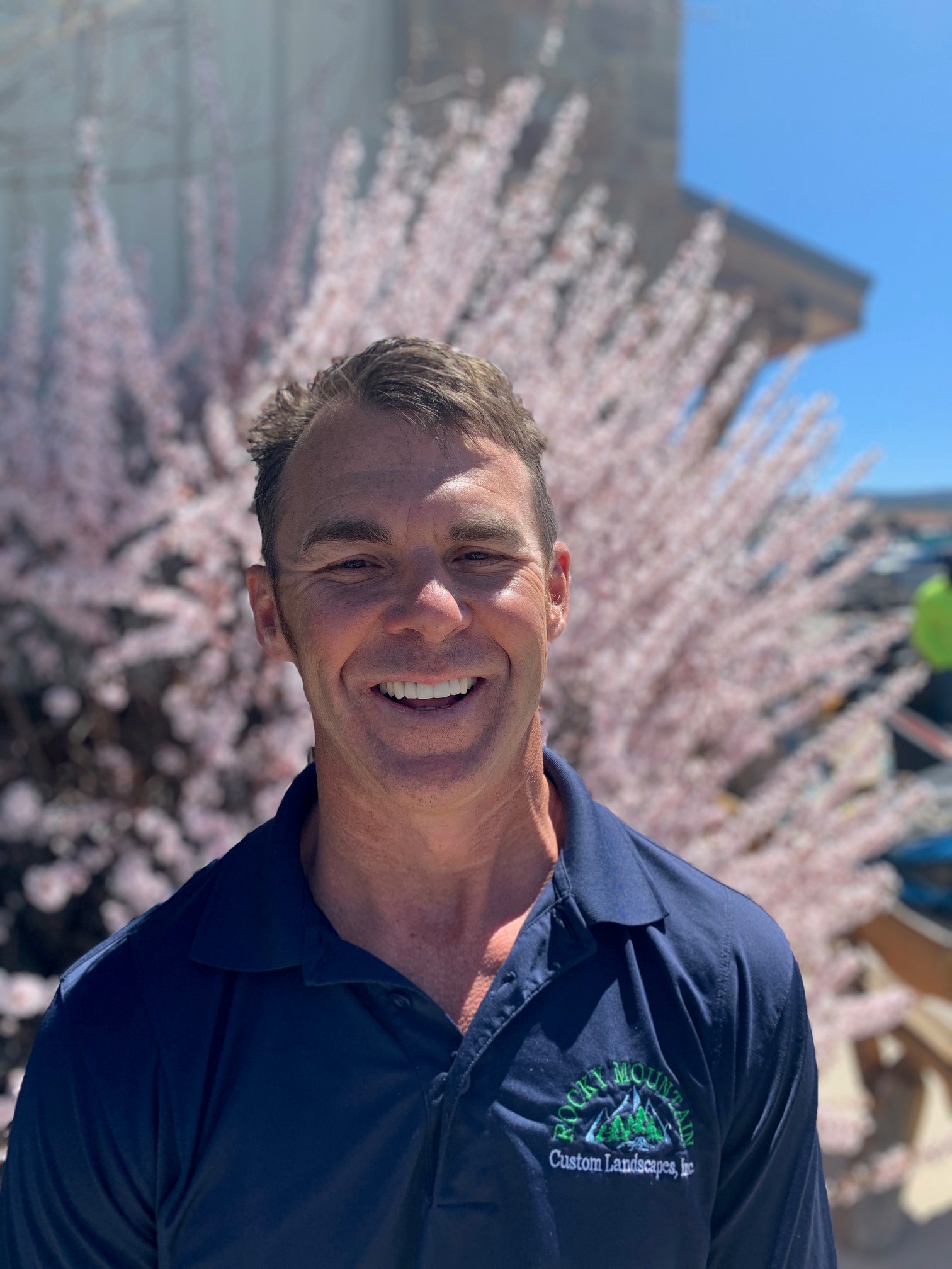 A smiling person stands in front of pink-blossomed trees, wearing a dark polo with an embroidered logo, on a sunny day.