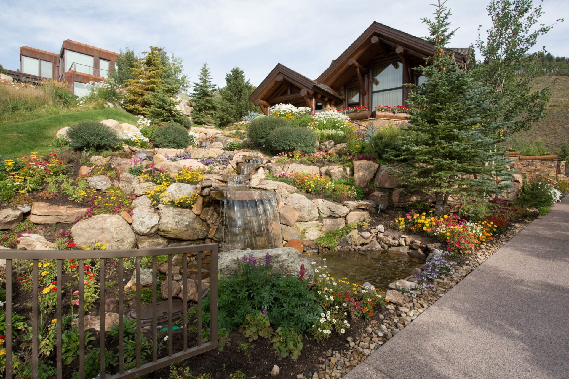 A beautifully landscaped garden with colorful flowers, a rock waterfall, and a pond in front of a house with large windows and a wooden facade.