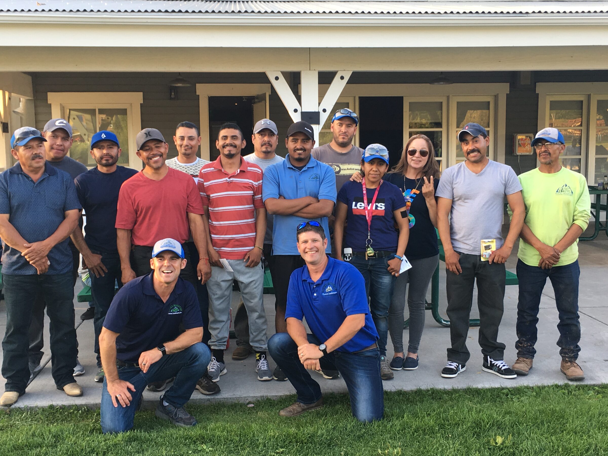 A group of people, possibly co-workers, are posing together outdoors in front of a building with a porch. They appear cheerful and casual.
