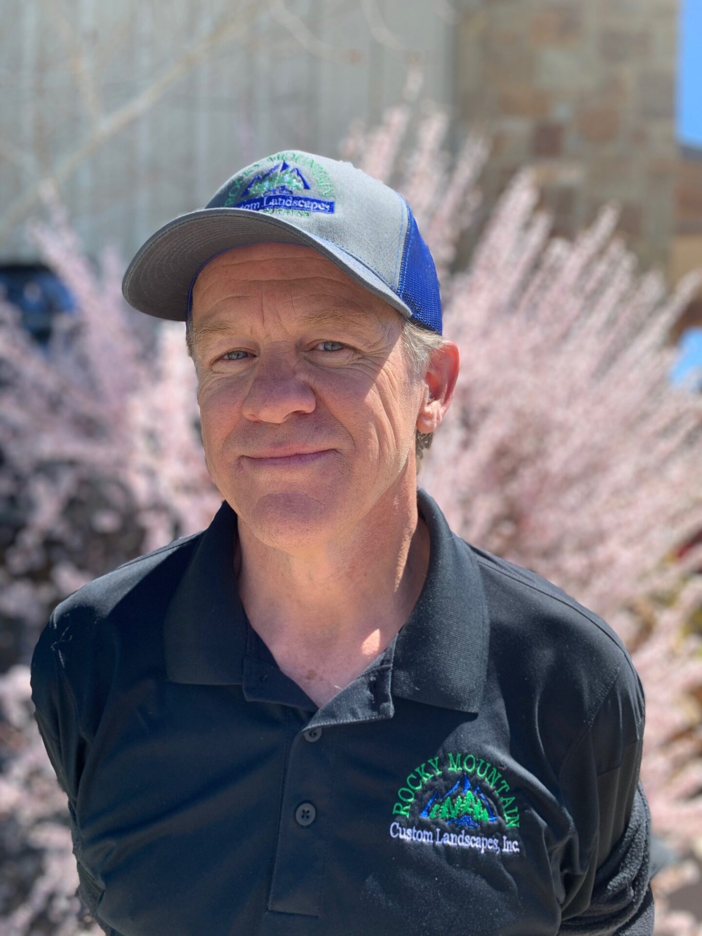 The image shows a person wearing a cap and a logo-embroidered polo shirt, standing before pink blooming bushes under a clear blue sky.