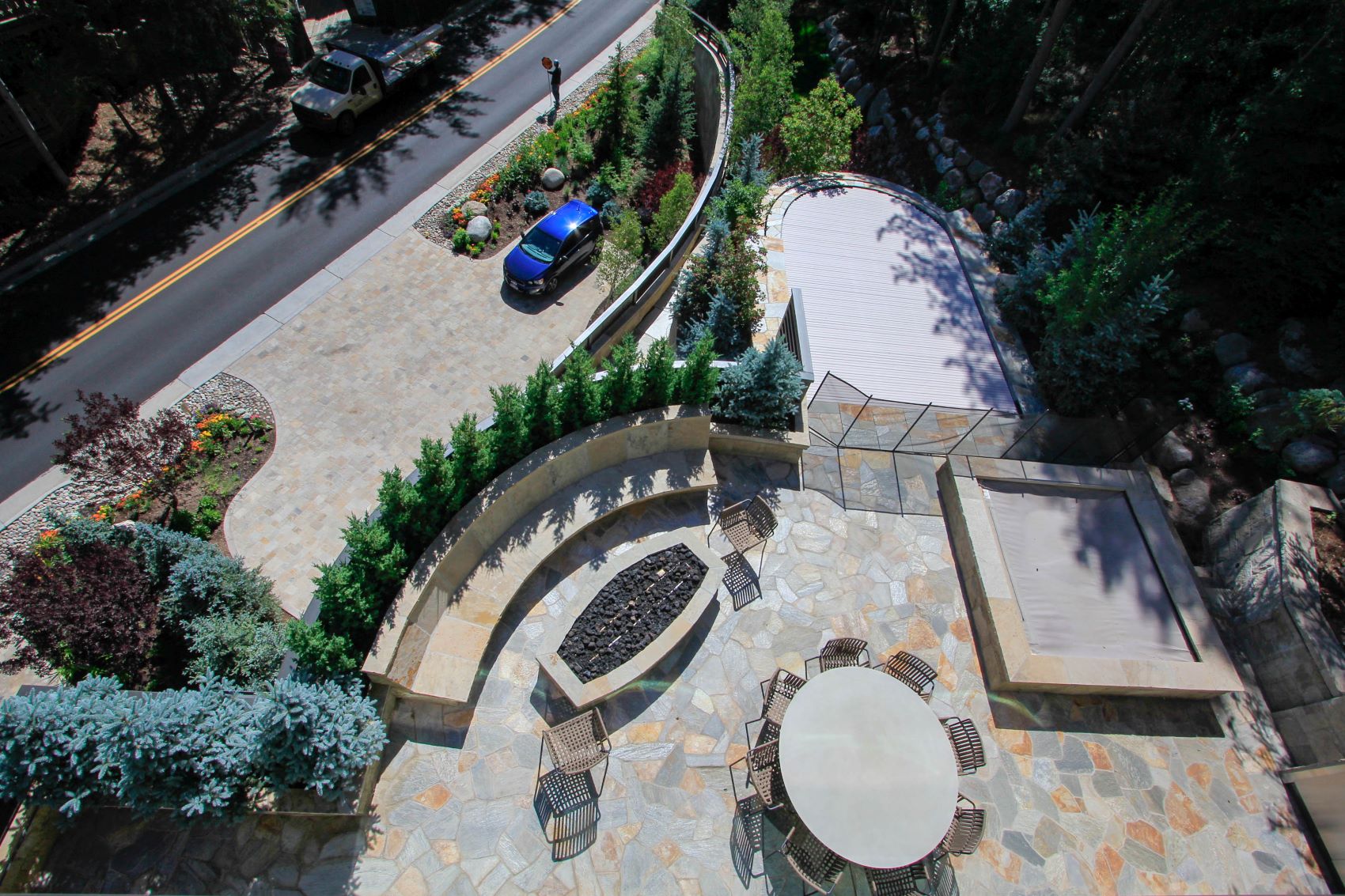 Aerial view of a landscaped terrace with a fire pit, seating area, and a road with a blue car and a person walking nearby.