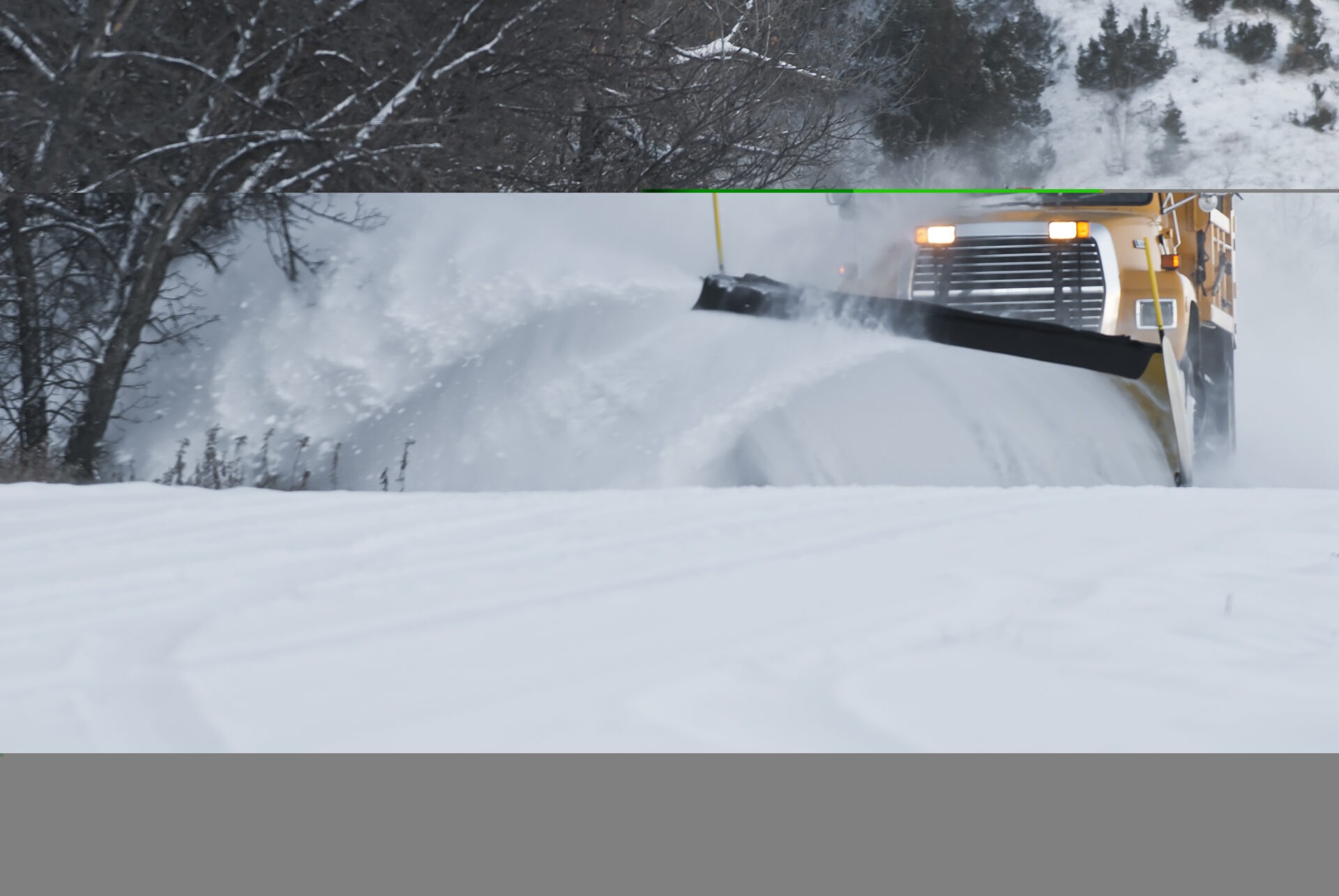 A snowplow is clearing a road, forcefully pushing snow aside, with trees in the background and visible snow cover on the ground.