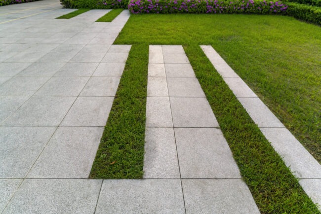 A walkway composed of alternating strips of concrete and grass, leading through a garden area with flowering shrubs, exhibiting a modern landscaping design.