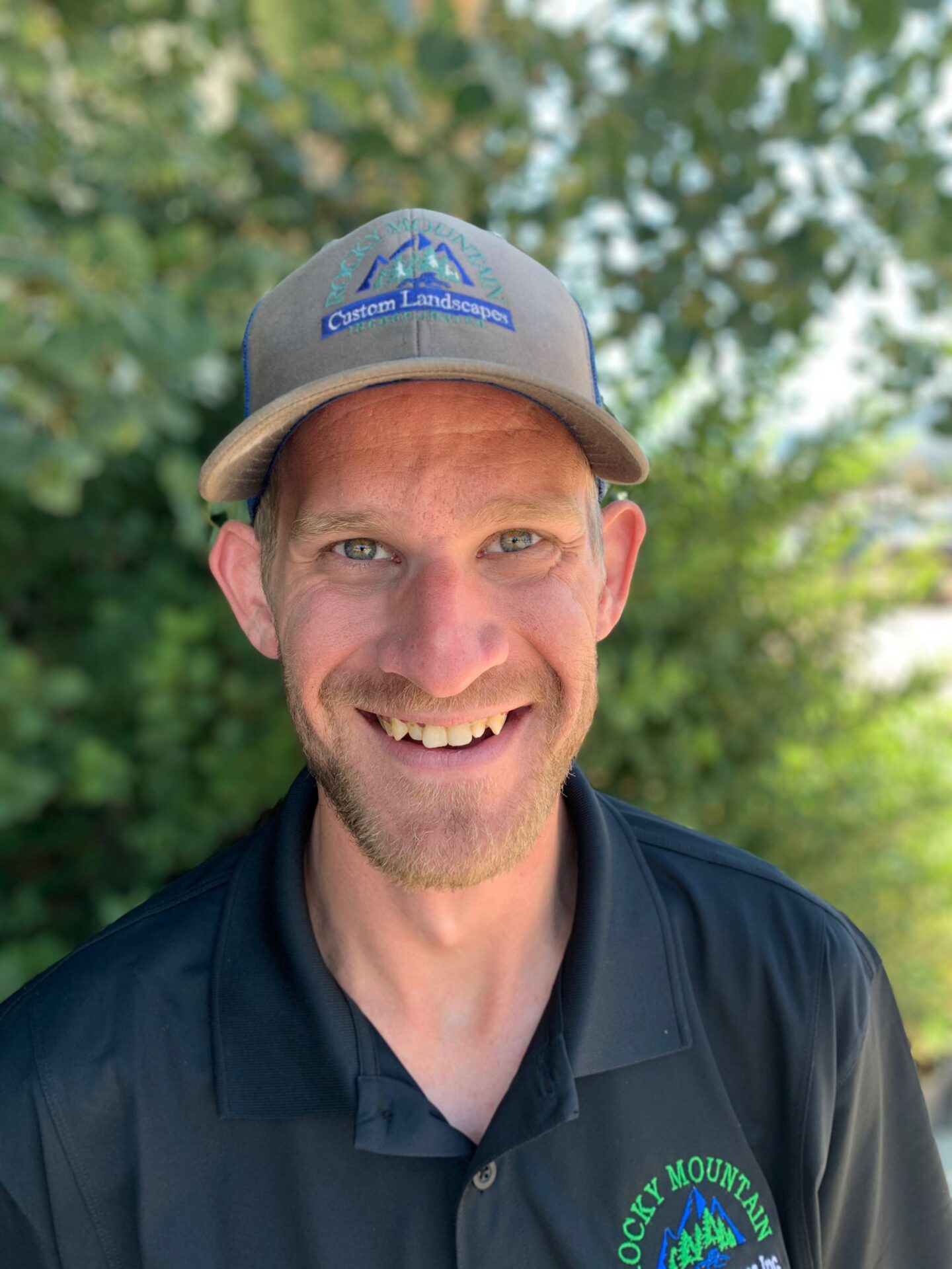 A person with a cap smiles at the camera against a blurred green backdrop. The cap's logo suggests a landscaping-related profession.