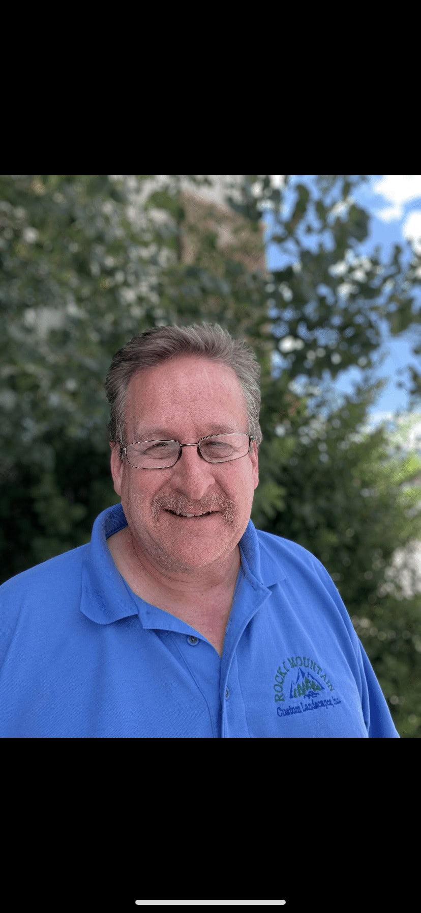 A person with glasses and a mustache smiles in front of blurred green foliage wearing a blue polo shirt with a company logo.