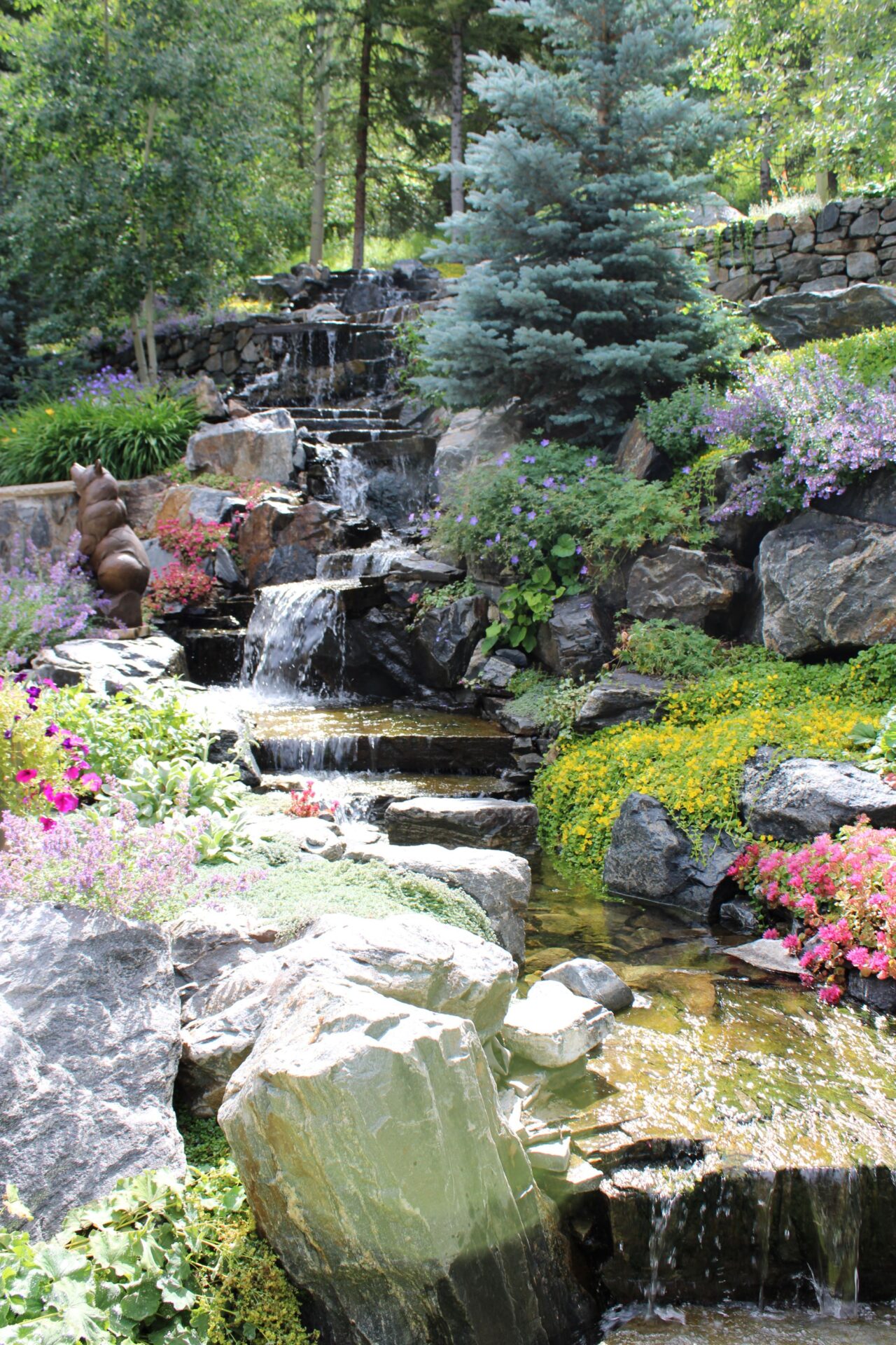 A landscaped garden with a cascading waterfall surrounded by colorful flowers, lush plants, and large rocks, under a sunny sky with trees.
