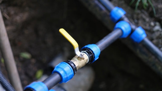 A yellow-handled brass ball valve is connected to a blue pipe, set against an unfocused background of natural ground and vegetation.