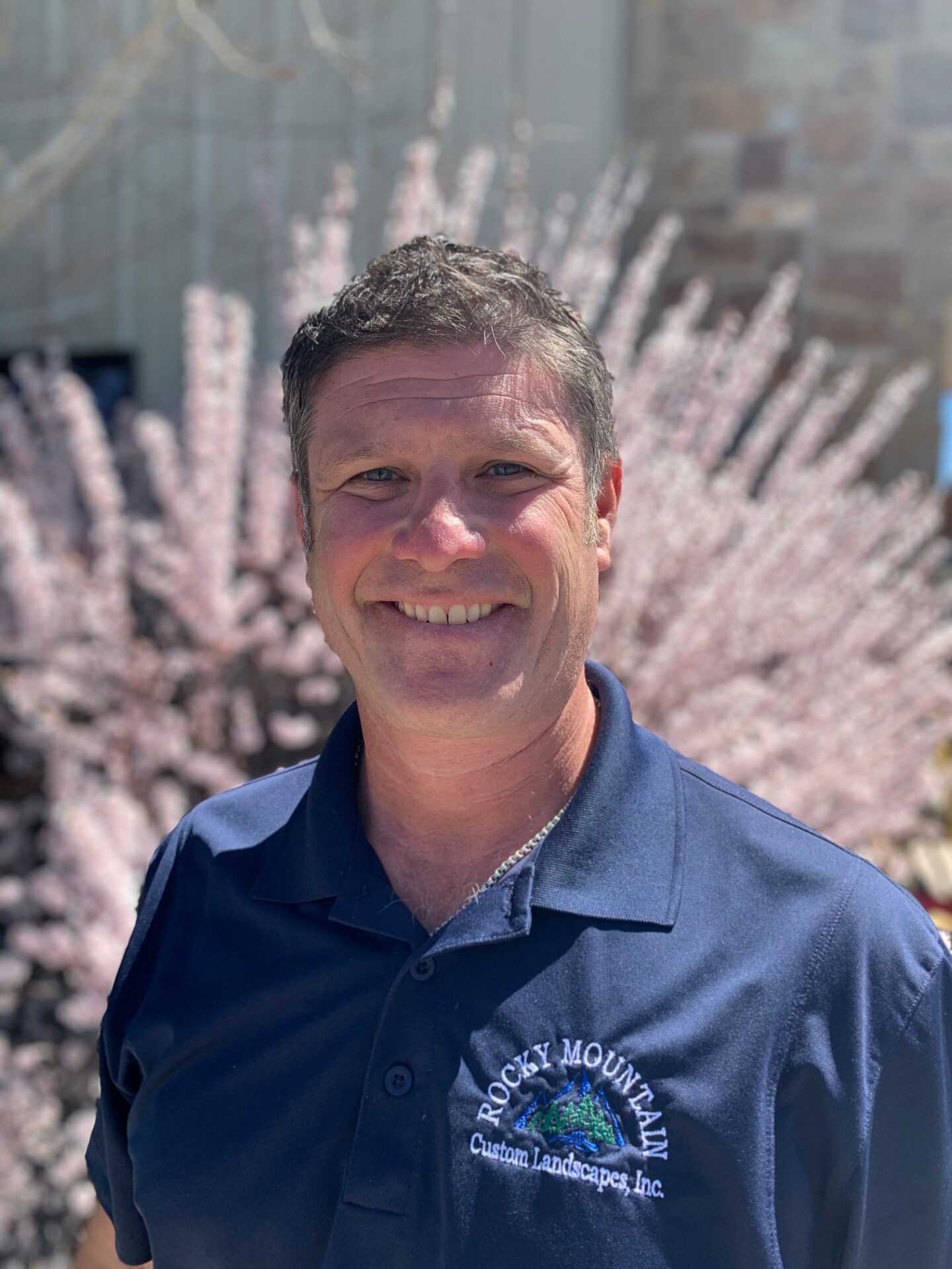 A smiling person stands outdoors in a blue uniform shirt with an embroidered logo, against a blurred background of pink cherry blossoms.