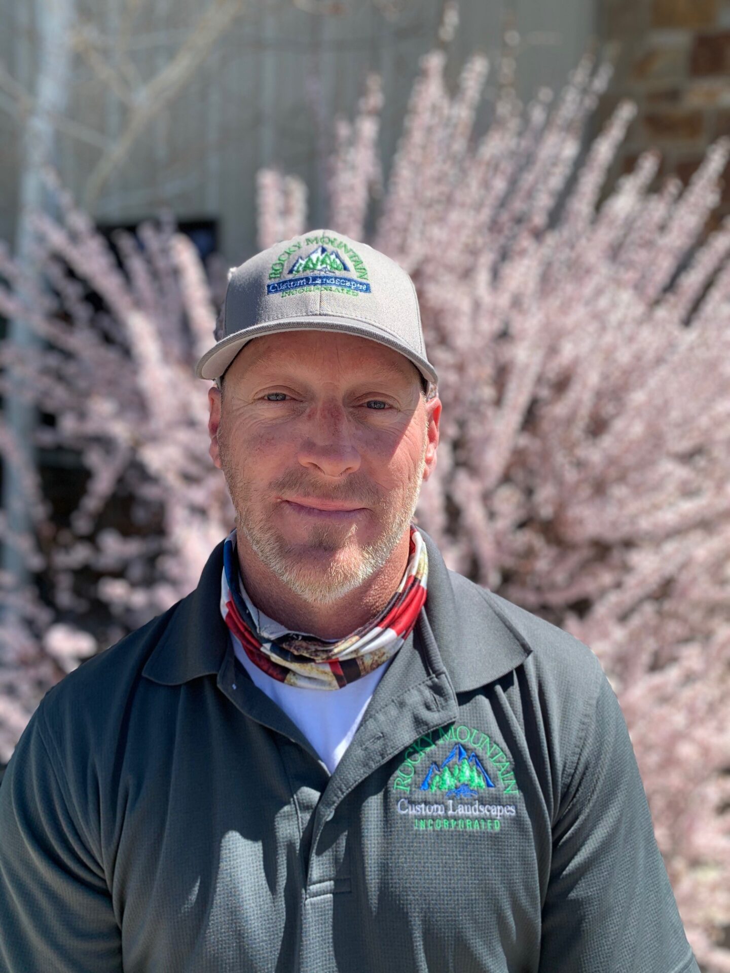 A person wearing a cap and work shirt stands in front of pink-blossoming trees, with a blurred background, in bright daylight.