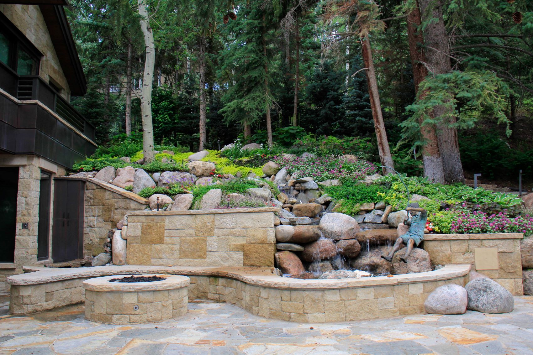 This image features a landscaped garden with a stone fire pit, a bench, and a small waterfall amidst boulders and lush plants, next to a building.
