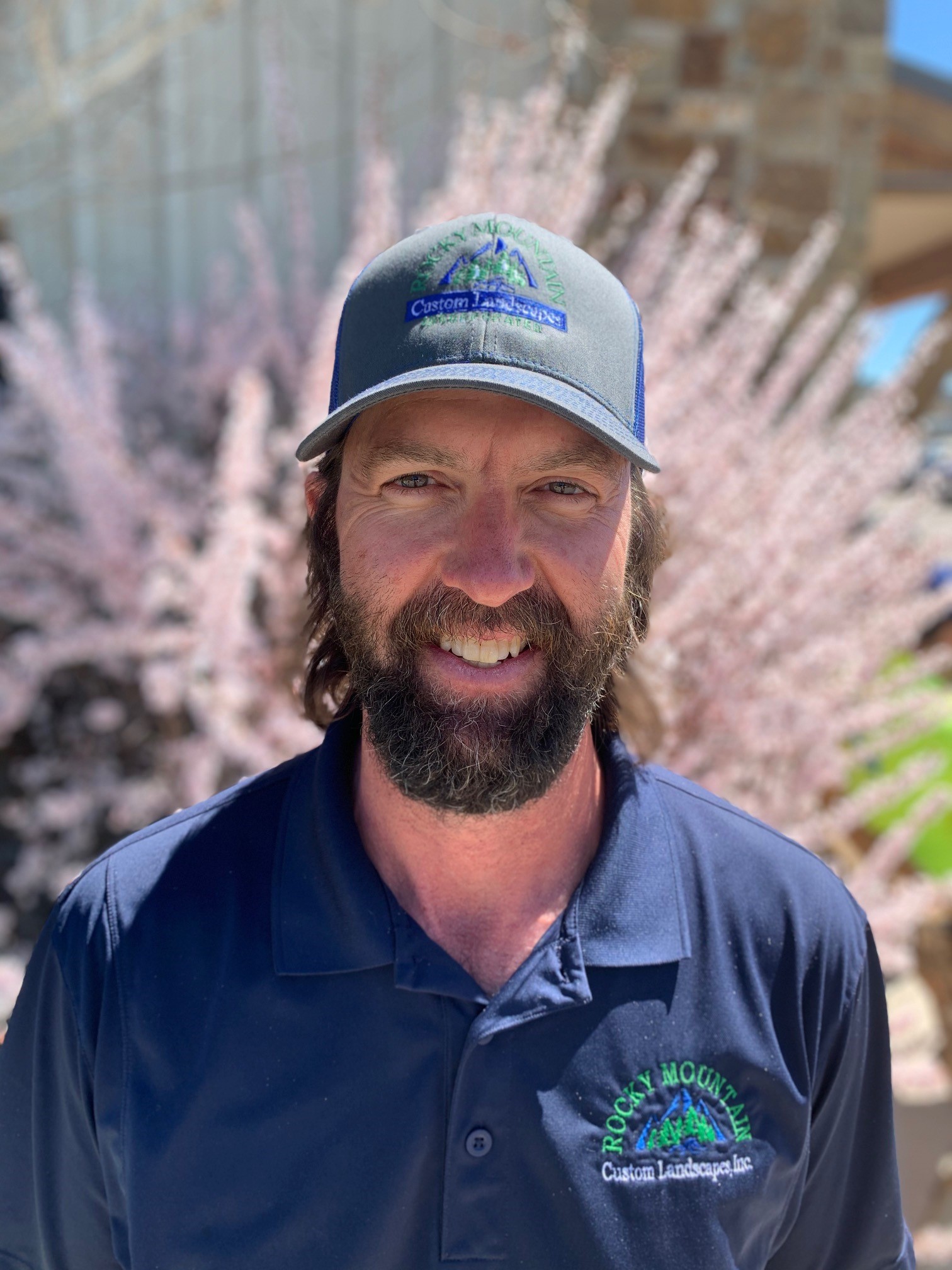 A smiling person with a beard wears a cap and a dark polo shirt embroidered with "Rocky Mountain Custom Landscape Inc." with flowering shrubs in the background.
