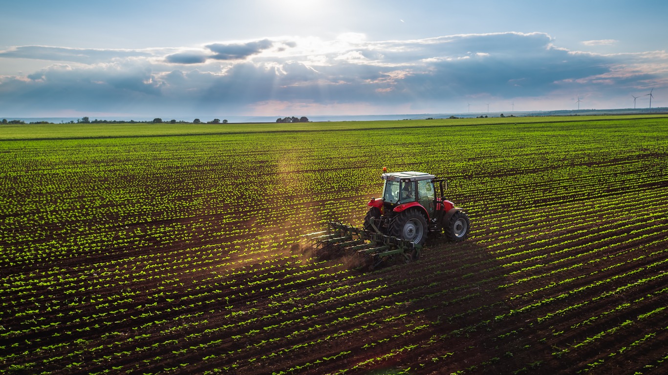 A tractor is working in a vast field with young crops under a sky punctured by sun rays, with distant wind turbines on the horizon.