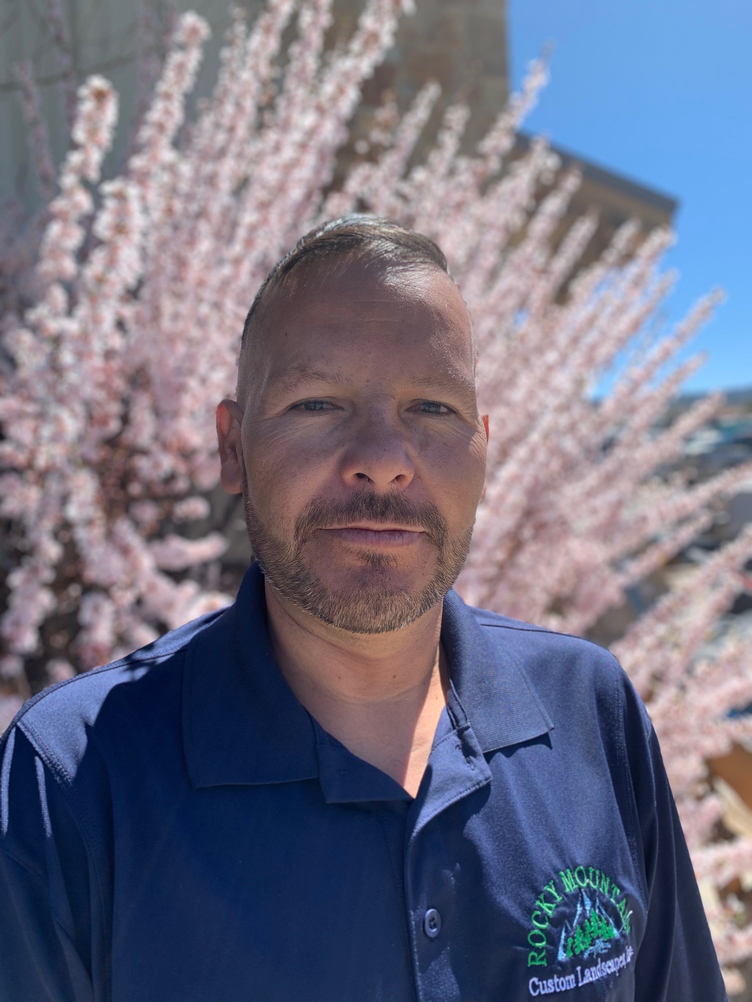 A person with short hair and a goatee is standing in front of pink blooming trees on a sunny day, wearing a blue shirt with an embroidered logo.