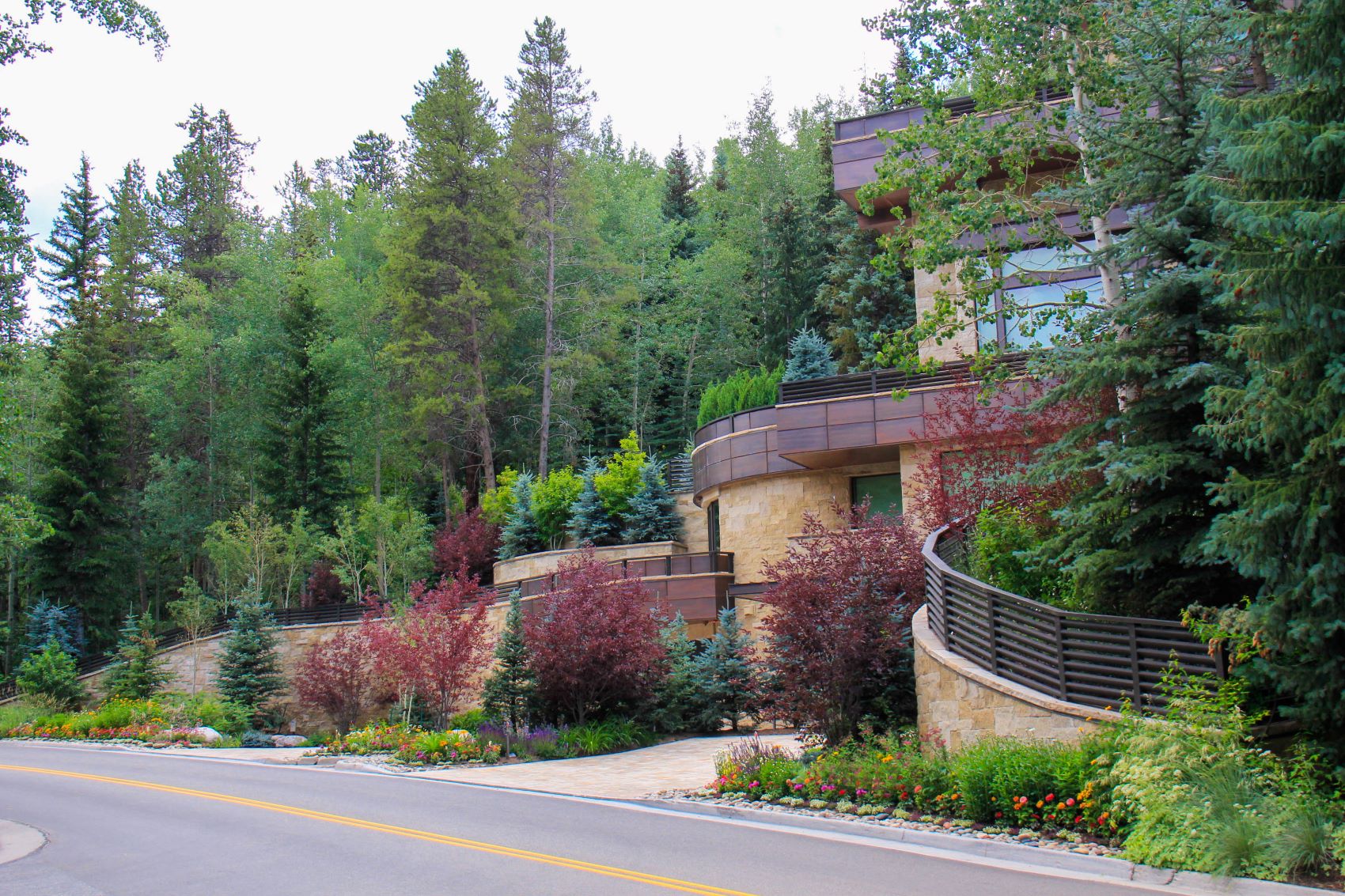 This image shows a modern building with curved architecture nestled among tall evergreen trees, with landscaped gardens along a paved road.