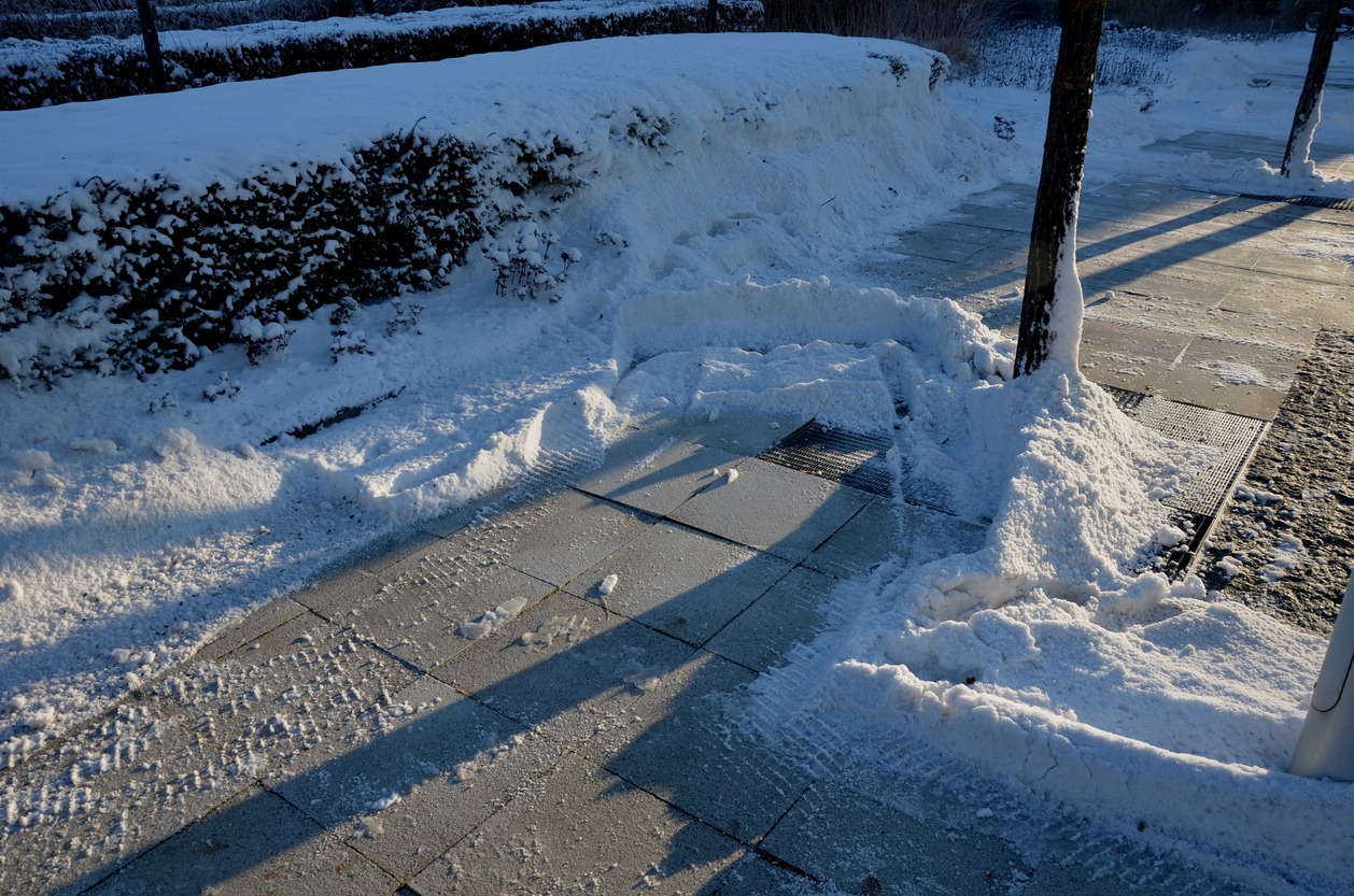 A snowy landscape with cleared sidewalk sections, snow piles, footprints, a shadow-casting tree, and sunlight reflecting off the icy surface.