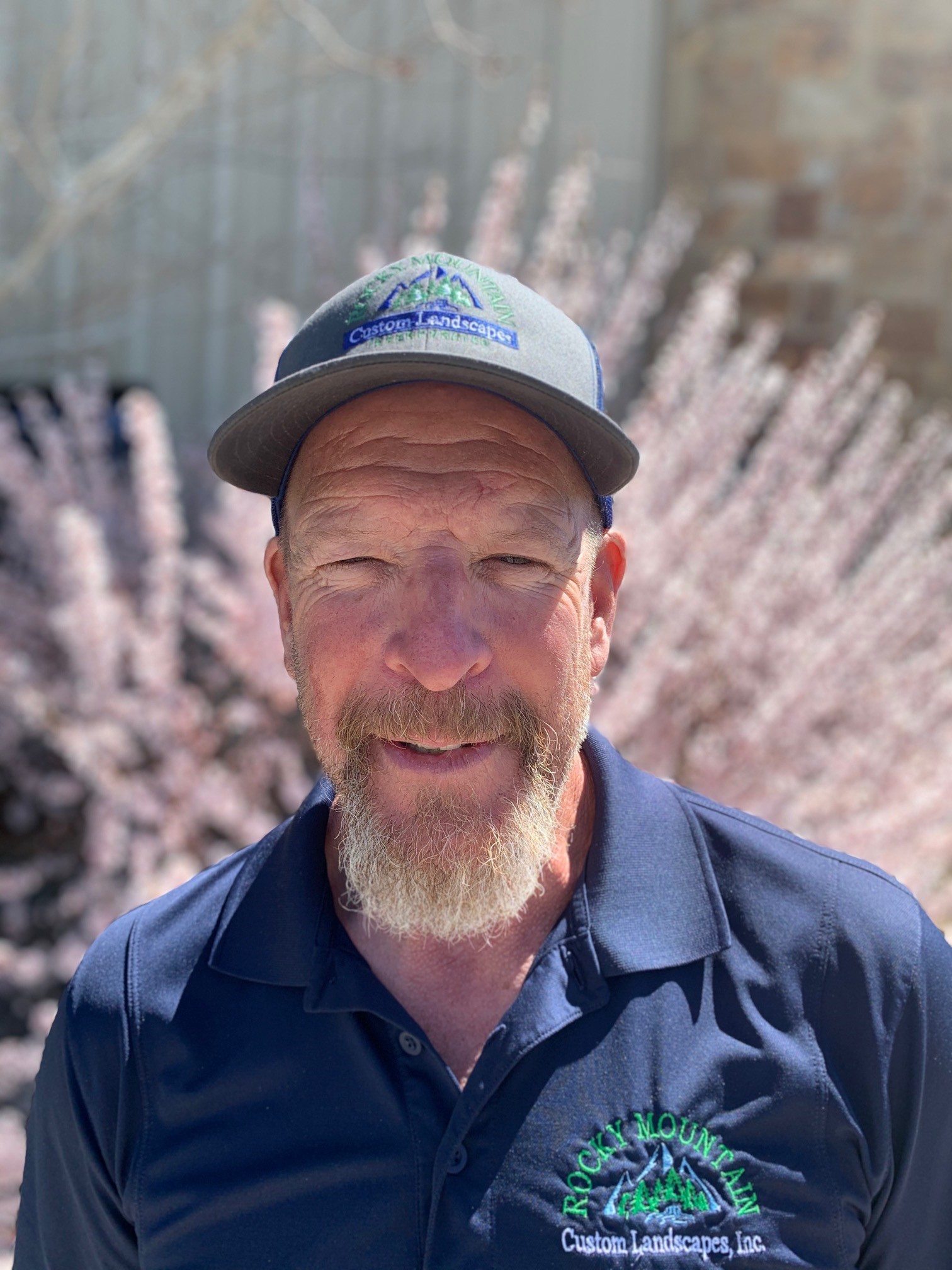 A smiling person wearing a cap and a polo shirt stands before blooming trees, showcasing a sunny day and a professional landscaping business logo.