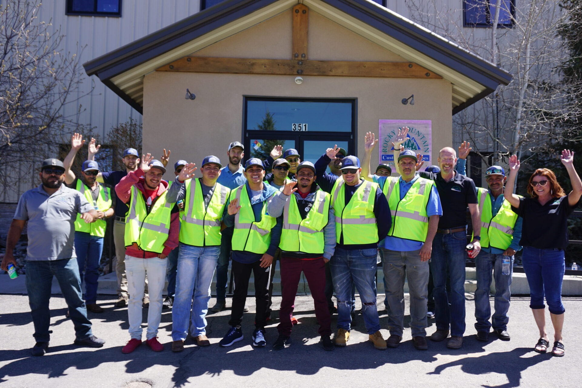 A group of happy people in reflective safety vests and caps posing in front of a building with trees and clear blue sky. Some are waving.