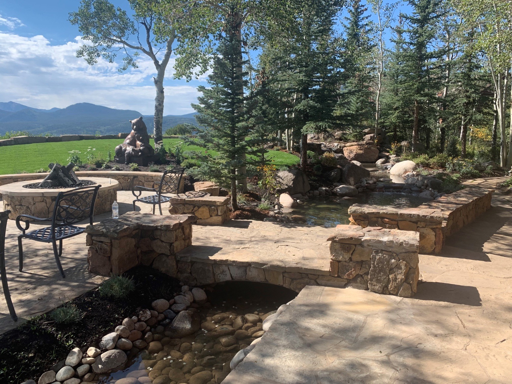 An outdoor landscaped area with stone structures, a water feature, fire pit, seating, and surrounding trees under a clear sky with distant mountains.