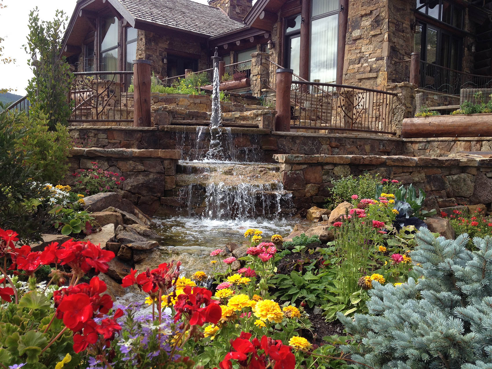 A rustic stone house with wooden beams features a multi-tier waterfall in a landscaped garden full of colorful flowers, including reds, yellows, and purples.