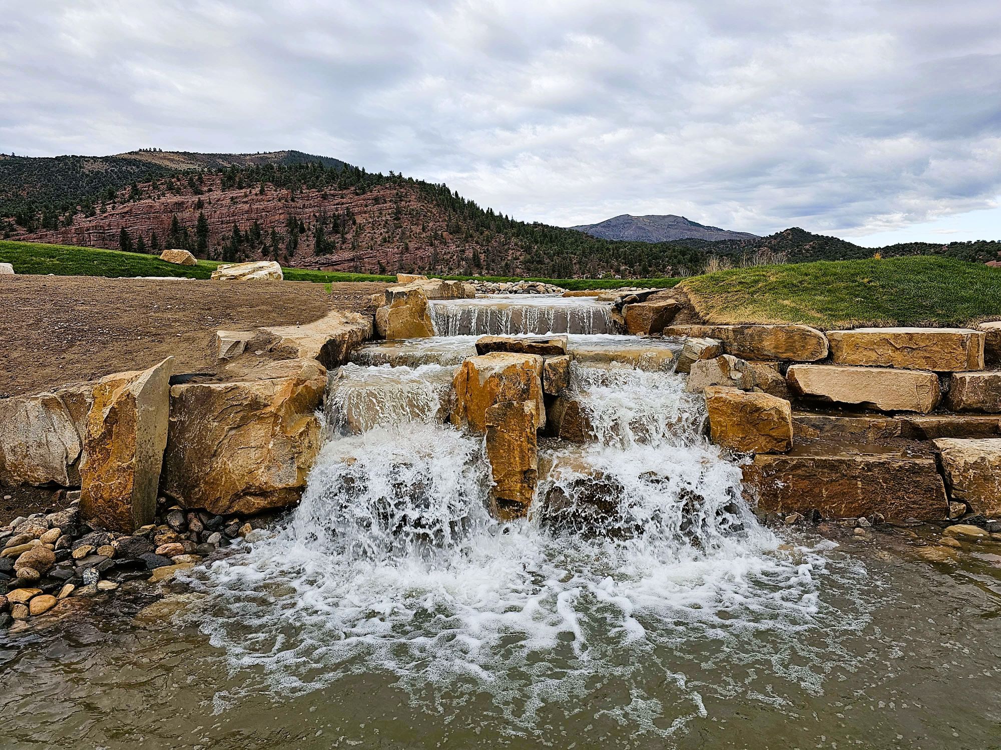 A small, man-made cascading waterfall with large rectangular rocks set against a backdrop of greenery and reddish-brown hills under a cloudy sky.