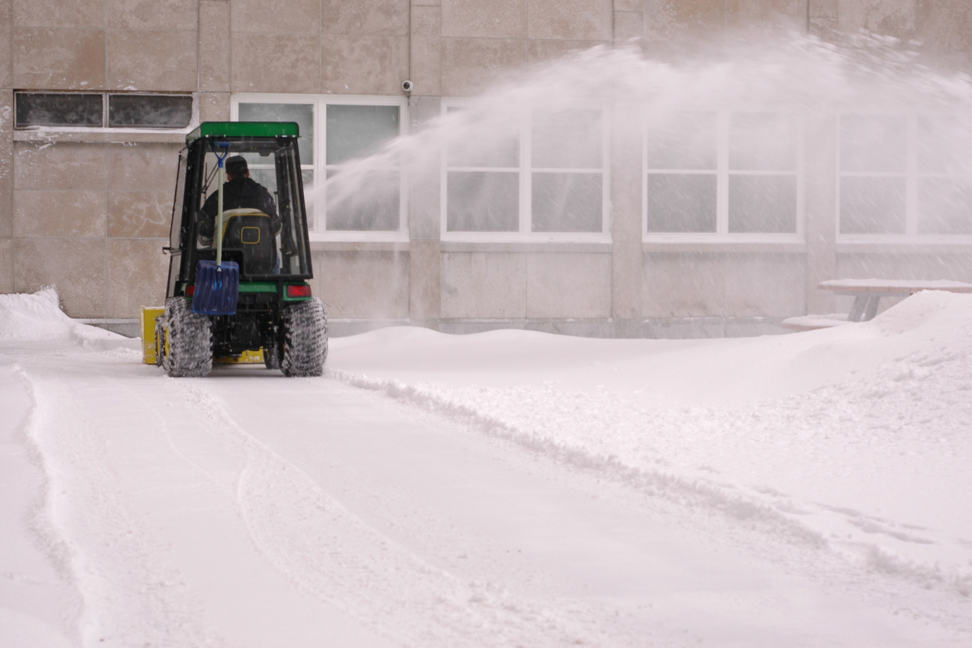 A person is operating a snowblower vehicle to clear snow near a building with large windows during winter, creating a spray of snow to the side.