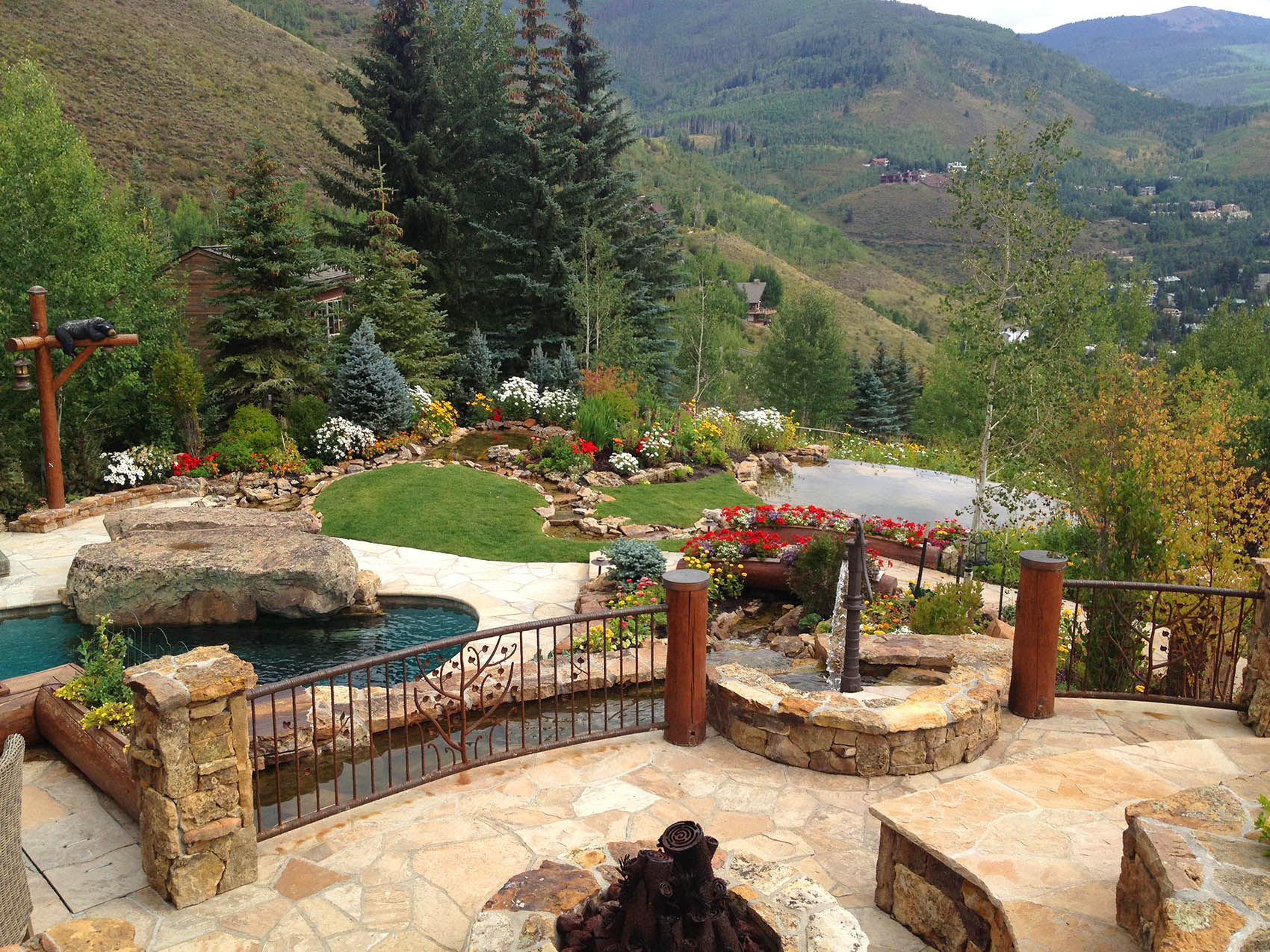 A luxurious backyard with a pool, hot tub, and lush landscaping overlooks a mountainous landscape. Stone pathways and vibrant flower beds enhance the scenic view.