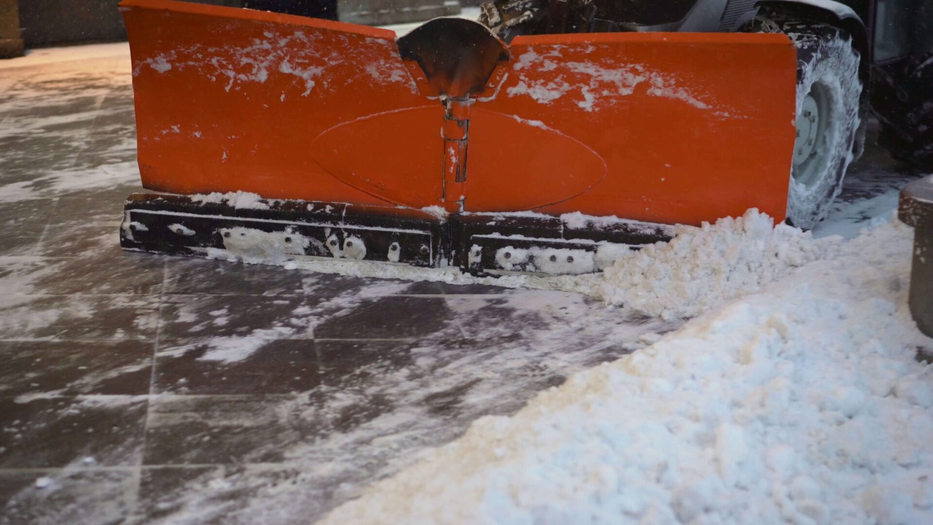 An orange snow plow attachment is clearing a snowy surface, pushing white snow to the side, with a vehicle partially visible behind it.