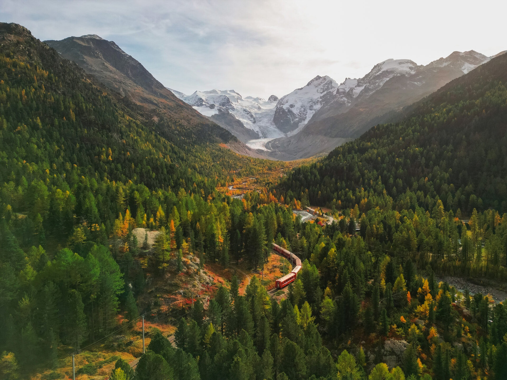 A scenic autumn valley with a red train running through it, surrounded by vibrant forests and snow-capped mountains under a soft light.