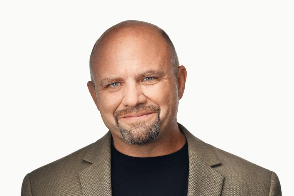 A person with a friendly smile, bald head, and stubble is pictured against a white background, wearing a black shirt and a brown blazer.