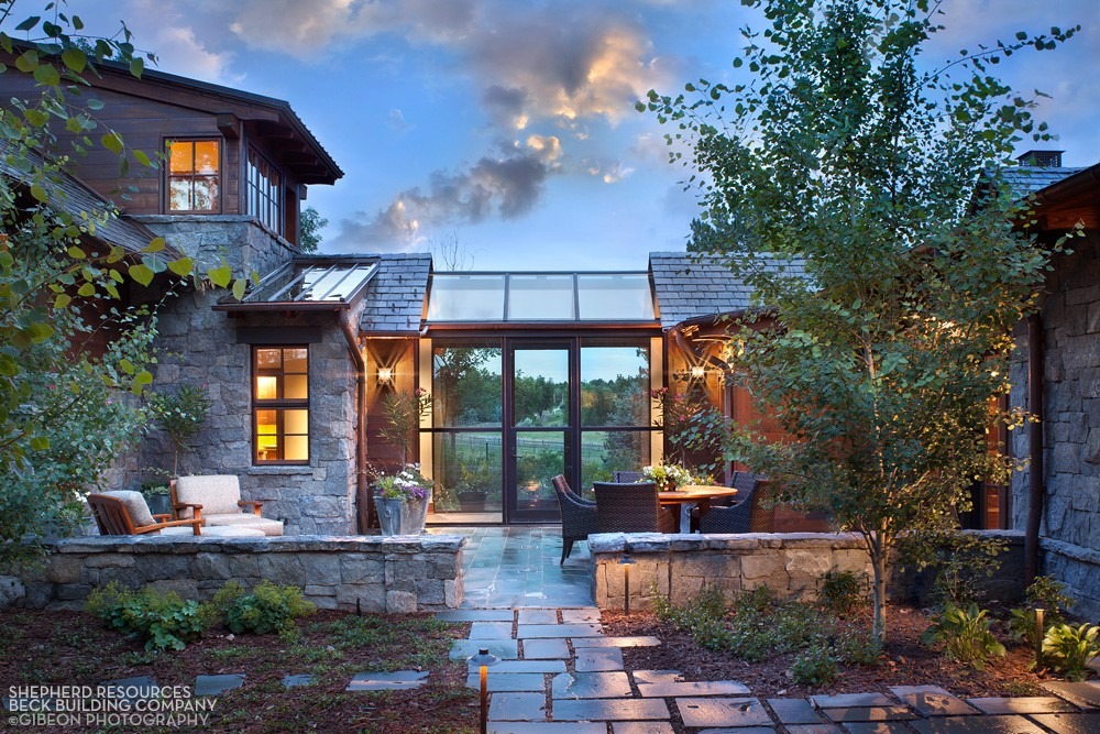 An elegant stone house at dusk with warm interior lights on, featuring large windows, outdoor seating areas, and lush greenery under a blue sky.
