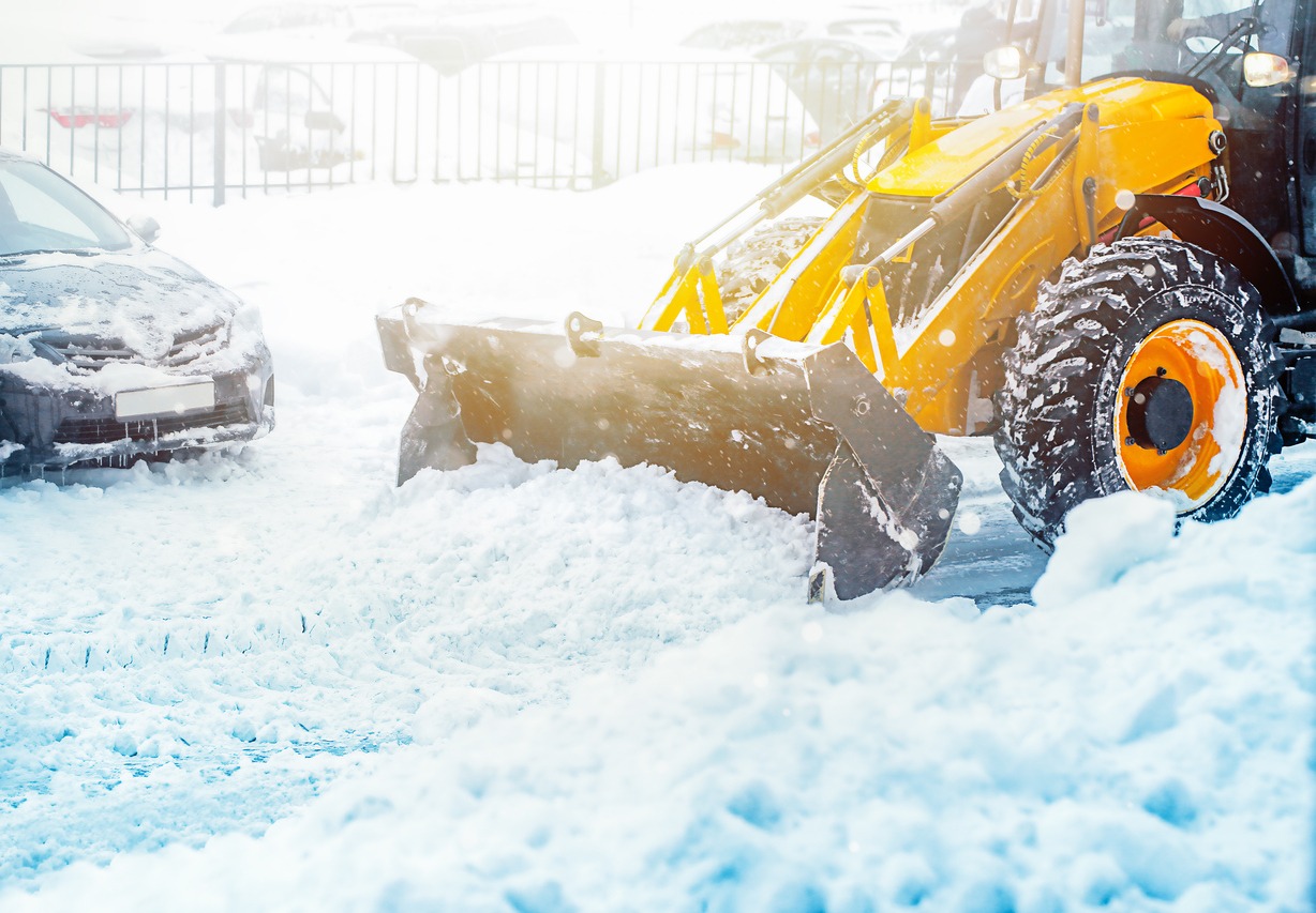 A yellow snowplow clears a snowy parking lot during a bright, wintry day, with a car partially covered in snow visible.