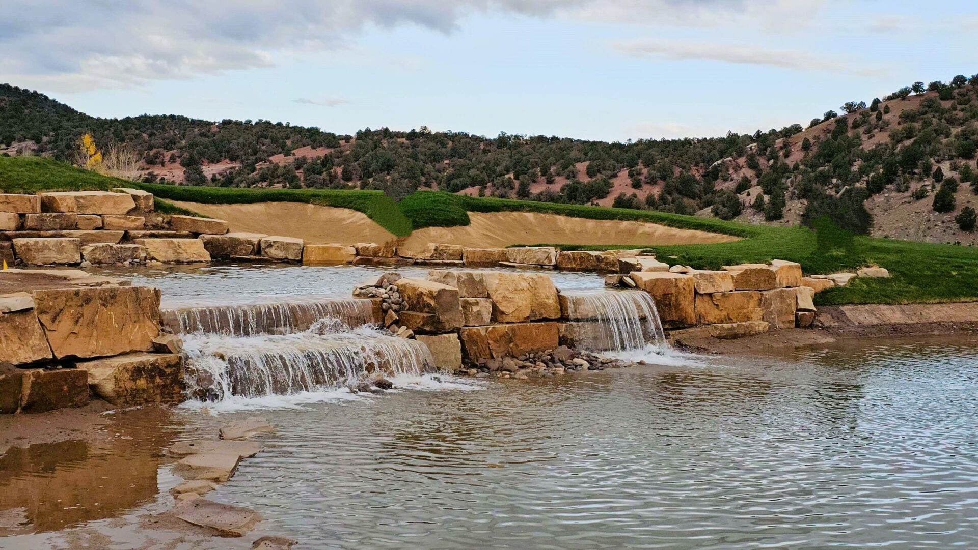This image shows a serene man-made waterfall cascading down large stone steps into a calm pond, surrounded by landscaped grass and rugged terrain.