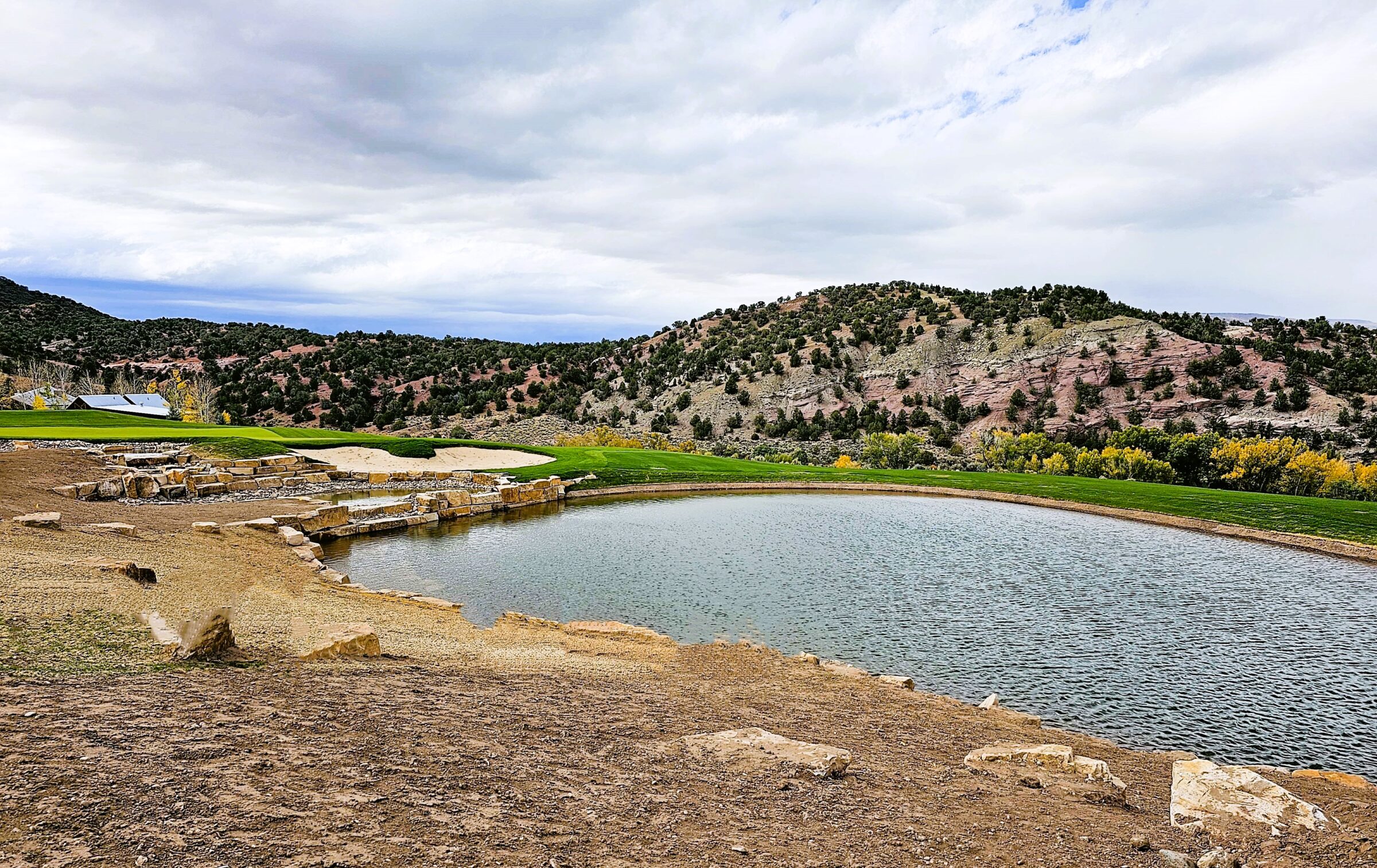 A tranquil man-made pond surrounded by stone steps, with green hills, trees, and a partly cloudy sky in the background. A building is partially visible.