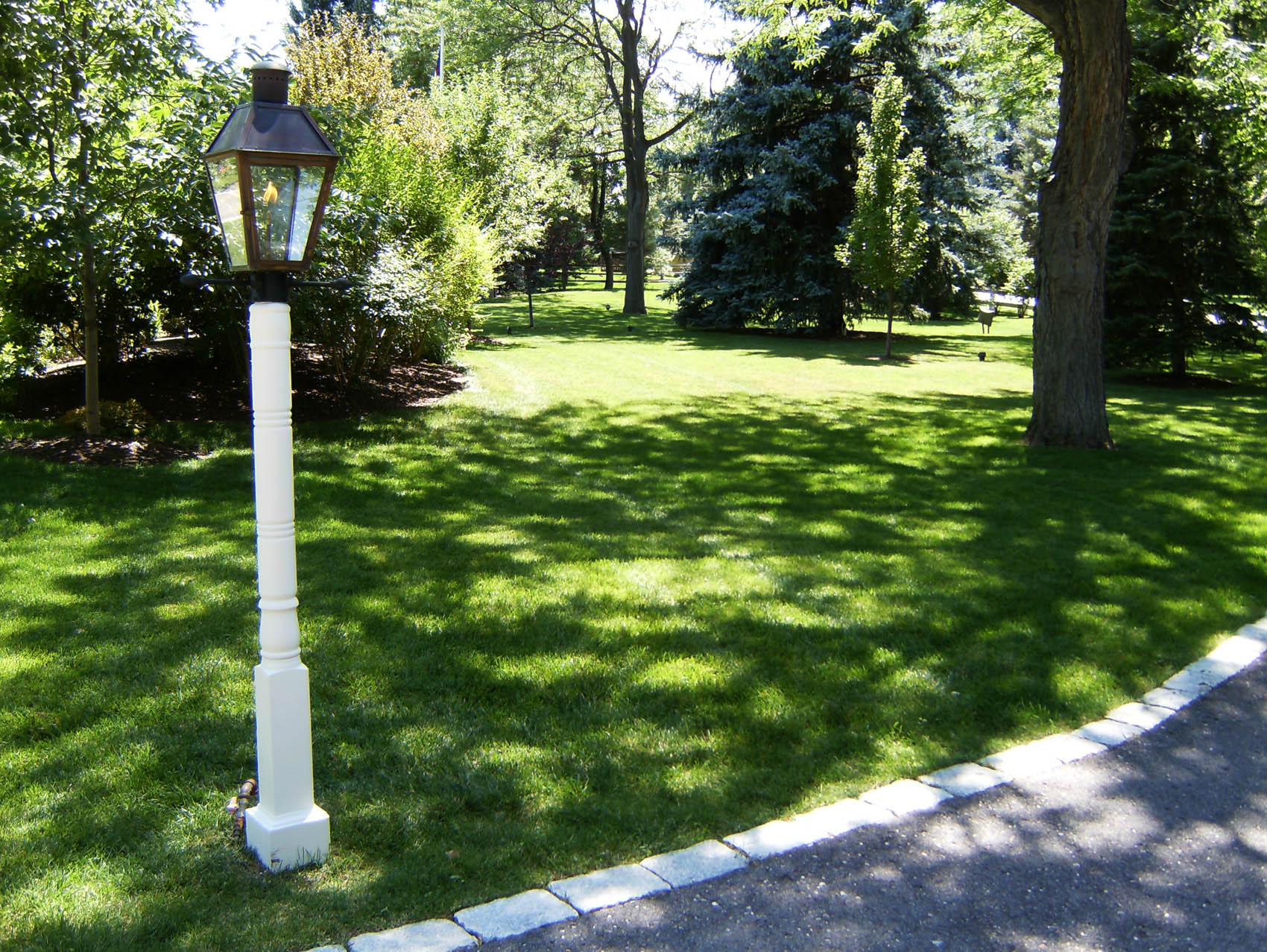An antique-style street lamp stands prominently in a lush, well-manicured park with green grass, trees, and a curved paved path lined with white stones.