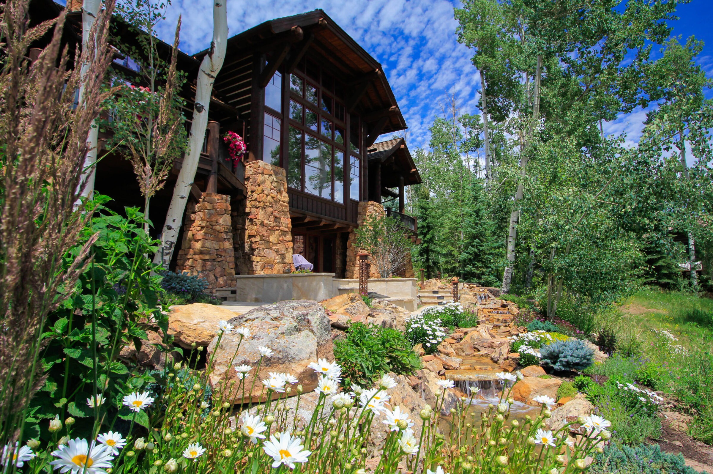 A large cabin with glass windows and stone pillars nestled in a green forest. A landscaped garden with flowers, rocks, and a small waterfall in the foreground.