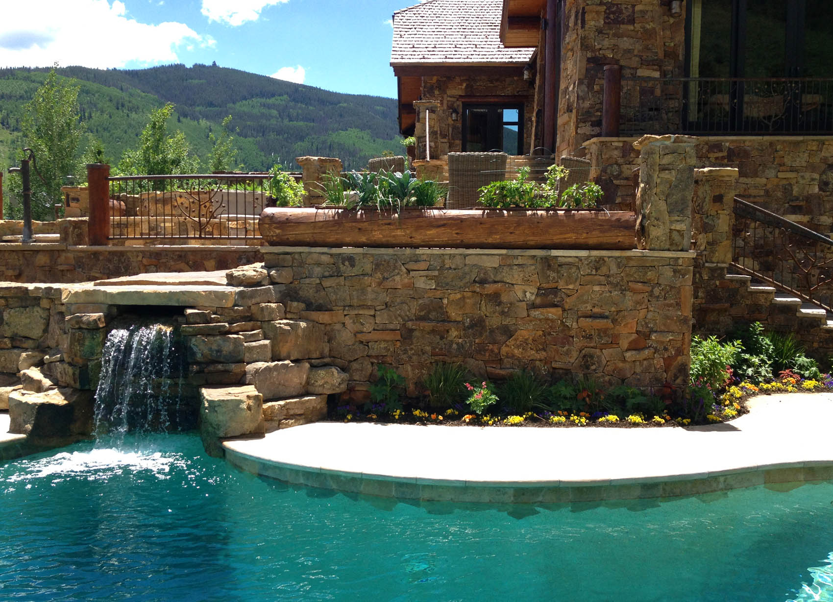 A luxurious outdoor pool with a stone waterfall feature beside a house with mountain views, surrounded by lush greenery and colorful flower beds.