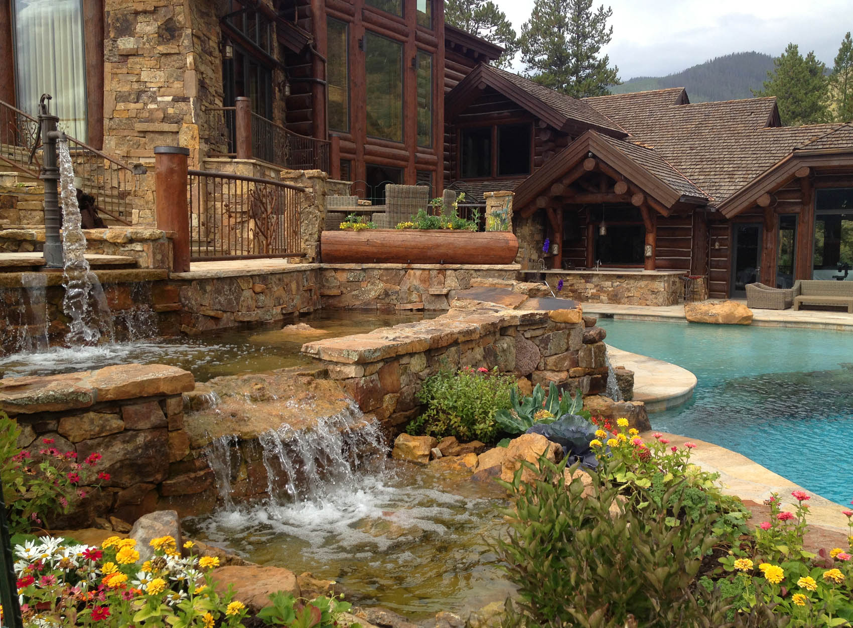 A luxurious backyard with a naturalistic swimming pool, stone waterfalls, colorful flowers, and a rustic wooden house with large windows and balconies.