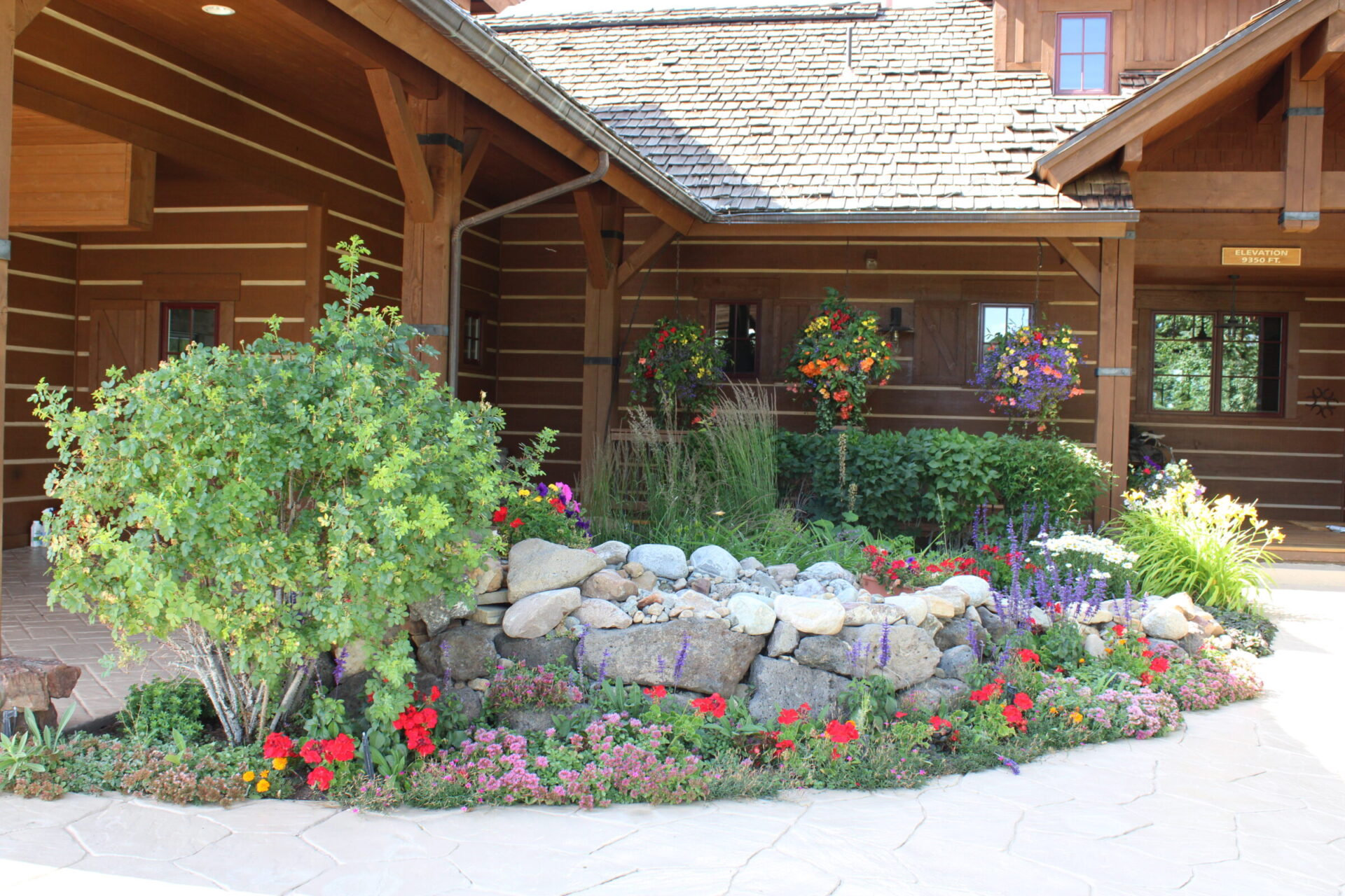 A well-maintained, colorful flowerbed in front of a wooden cabin, featuring various flowers, rock elements, and hanging flower pots under a clear sky.