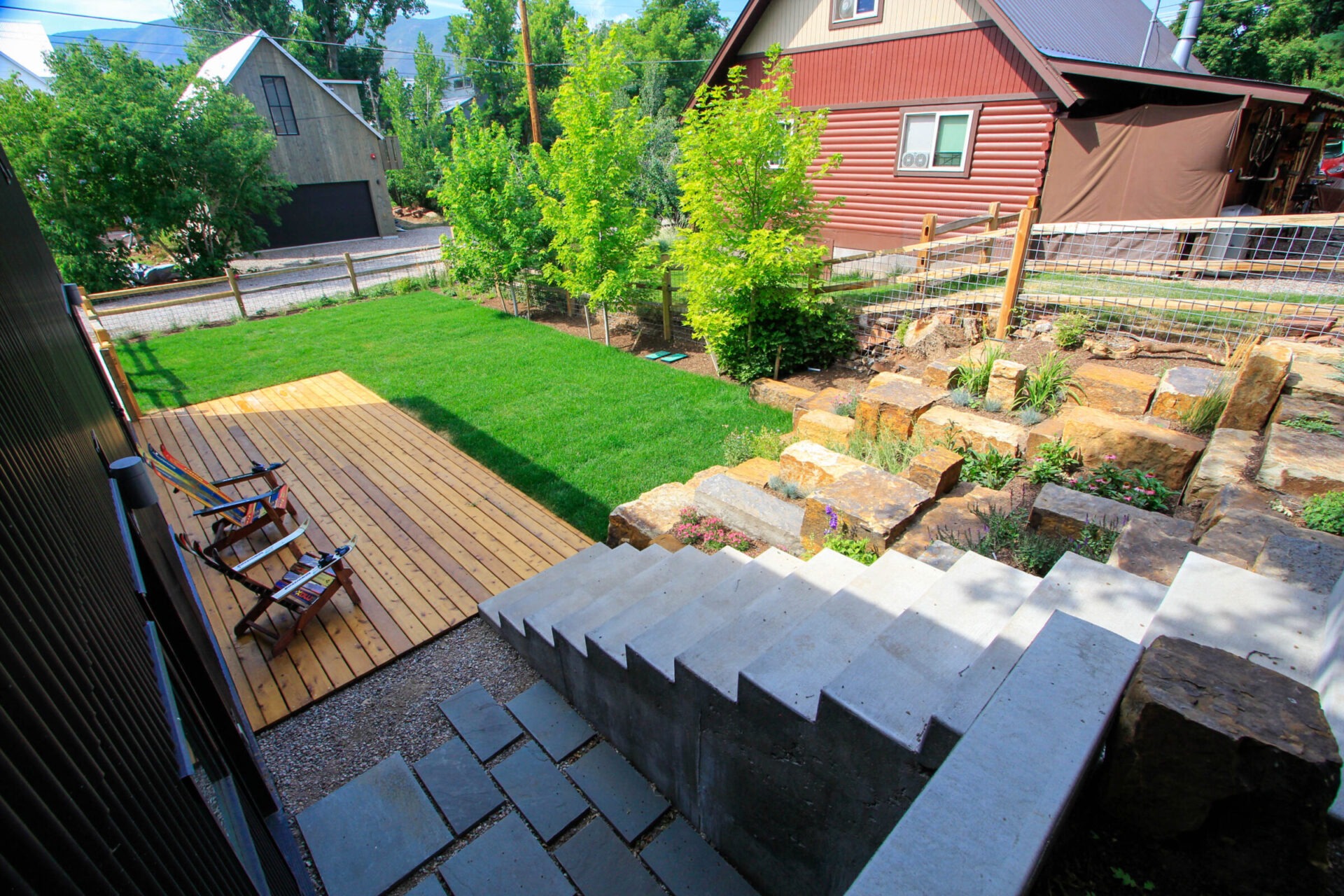 An elevated view of a terraced backyard with a wooden deck, concrete stairs, a lush lawn, landscaping with stone walls, and nearby buildings.