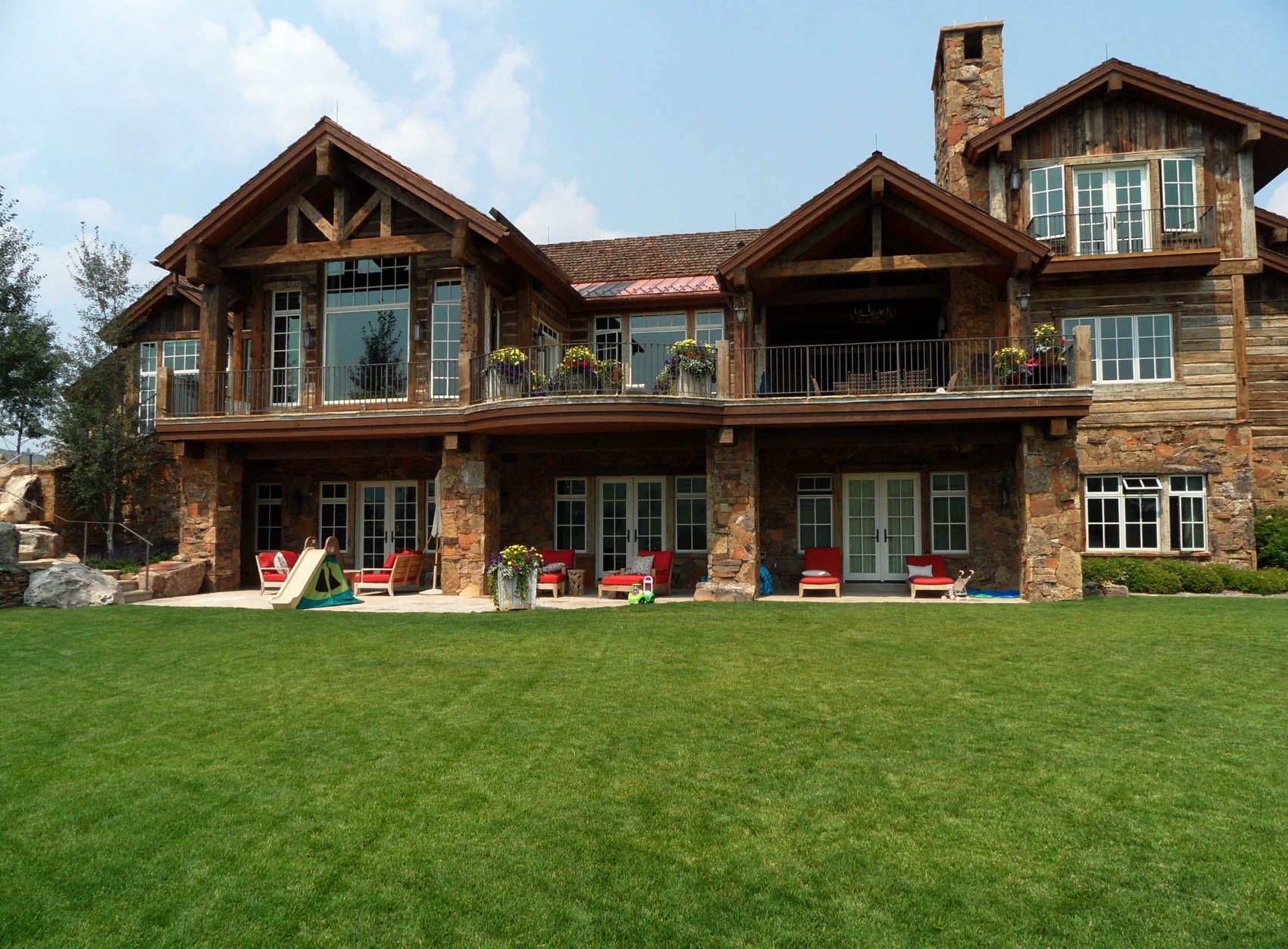 A large two-story house with stone and wood finish, balconies, and a well-manicured lawn with outdoor chairs and children's playthings.