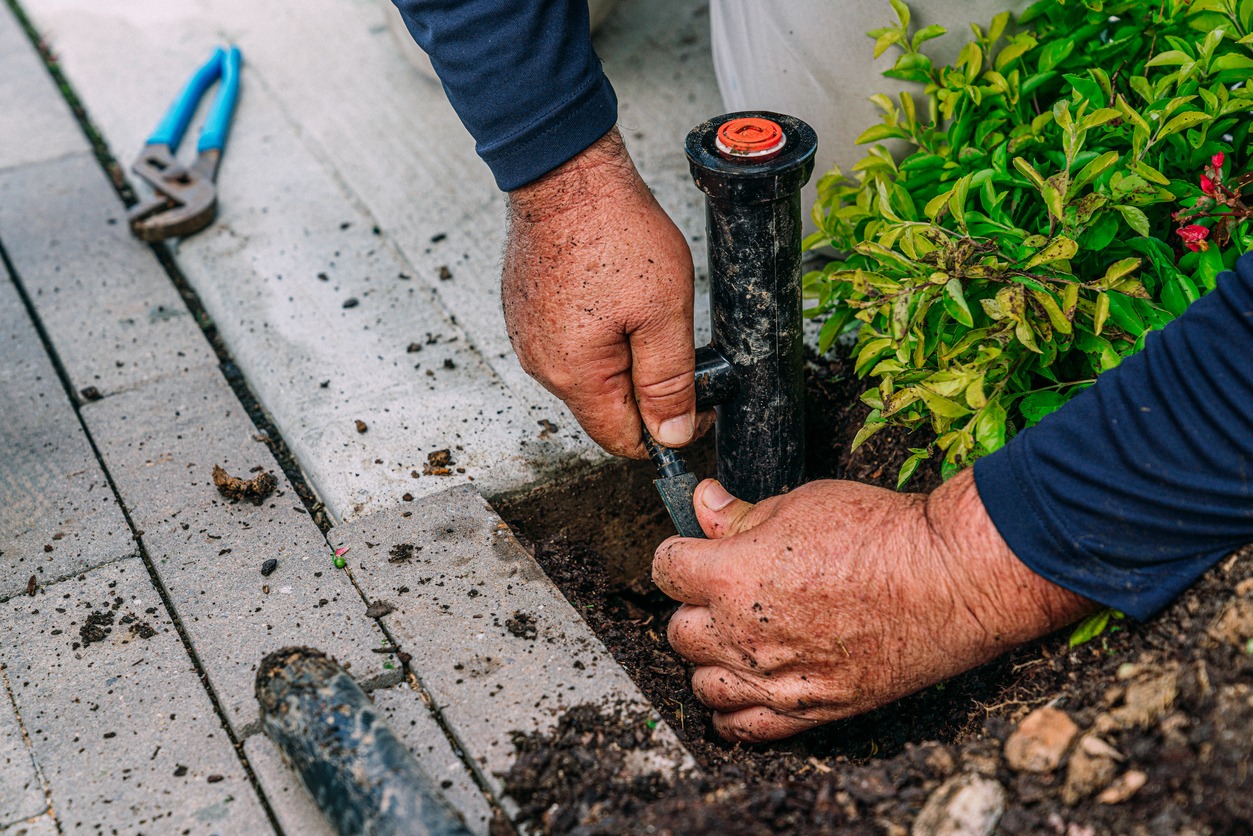 A person is installing or repairing an irrigation system by a pathway, with soil-covered hands, visible pliers, and lush greenery nearby.