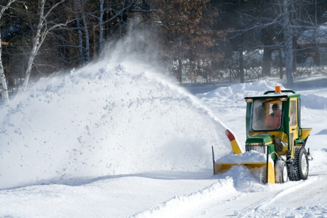 A person operates a green and yellow snowblower, ejecting a large arc of snow onto a snowbank under a clear blue sky with trees.