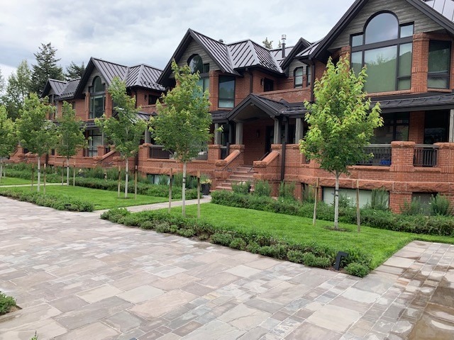 A row of red brick townhouses with dark roofs, each featuring a front porch. A neatly landscaped pathway with young trees leads up to the homes.